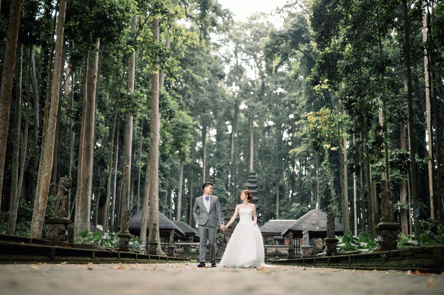 Bride and groom hold hands in a lush forest wedding photo with tall trees and a stone temple backdrop.