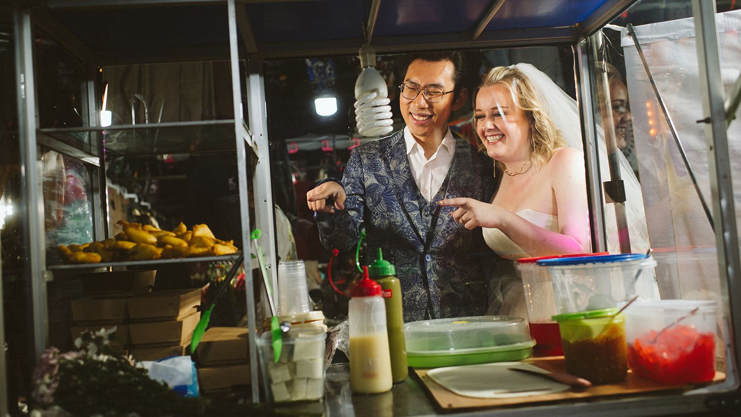 Couple smiling together at a vibrant night market food stall with colorful sauces and fresh produce.