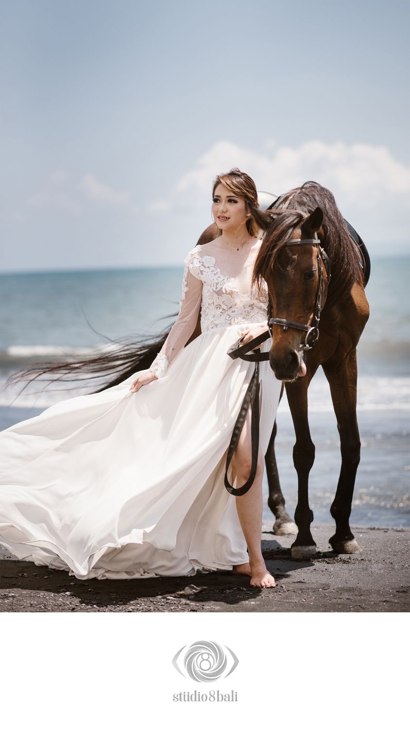 Bride in white lace gown stands beside a brown horse on a black sand beach with ocean backdrop.