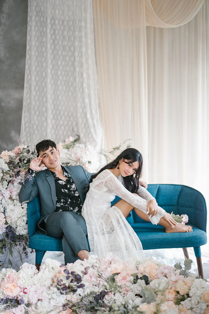 Couple posing on a teal sofa surrounded by pastel florals, woman in white lace dress, man in gray suit, elegant studio setting.