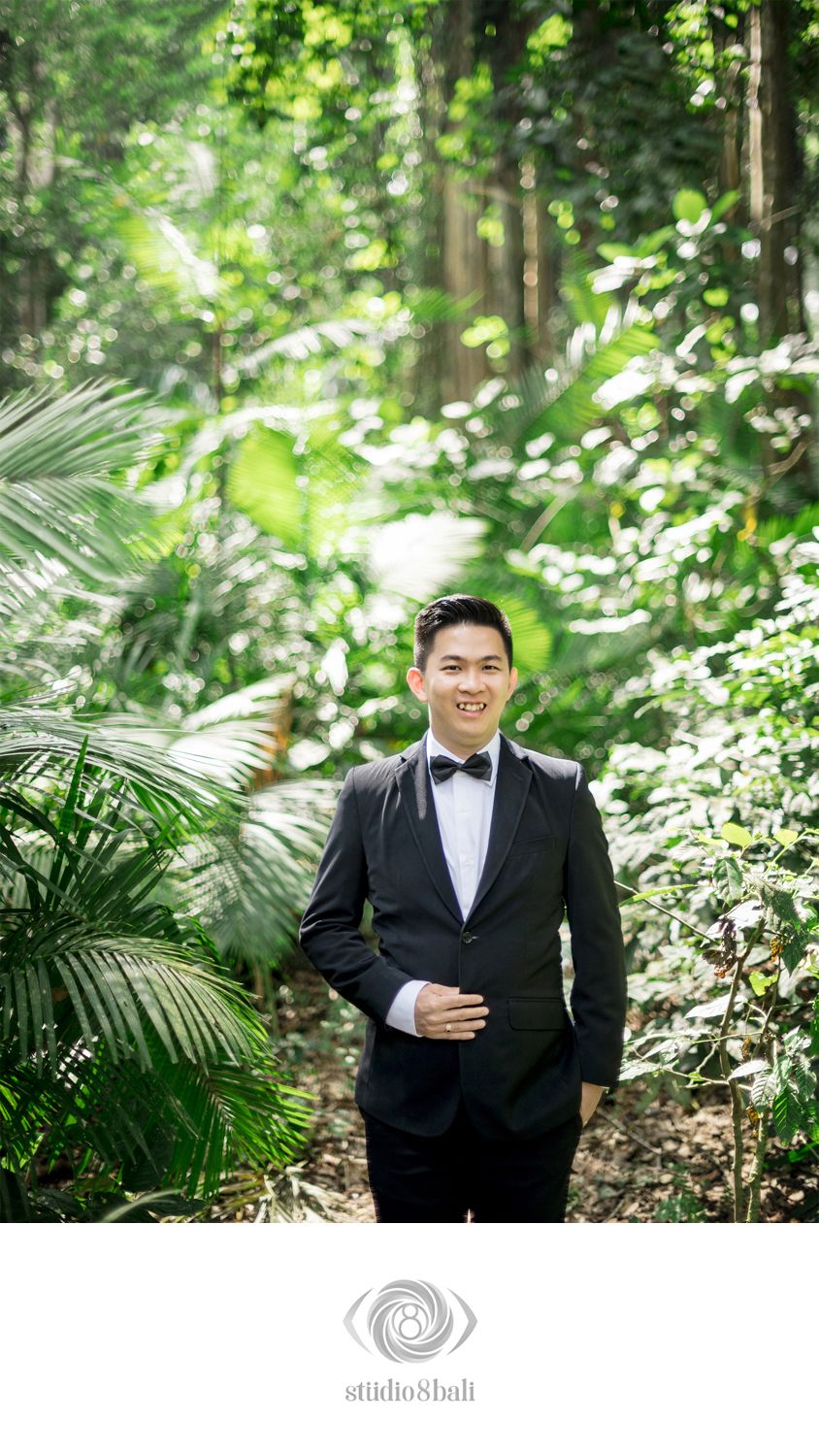 Groom in black tuxedo with bow tie smiling in lush tropical forest setting for Bali wedding photography.