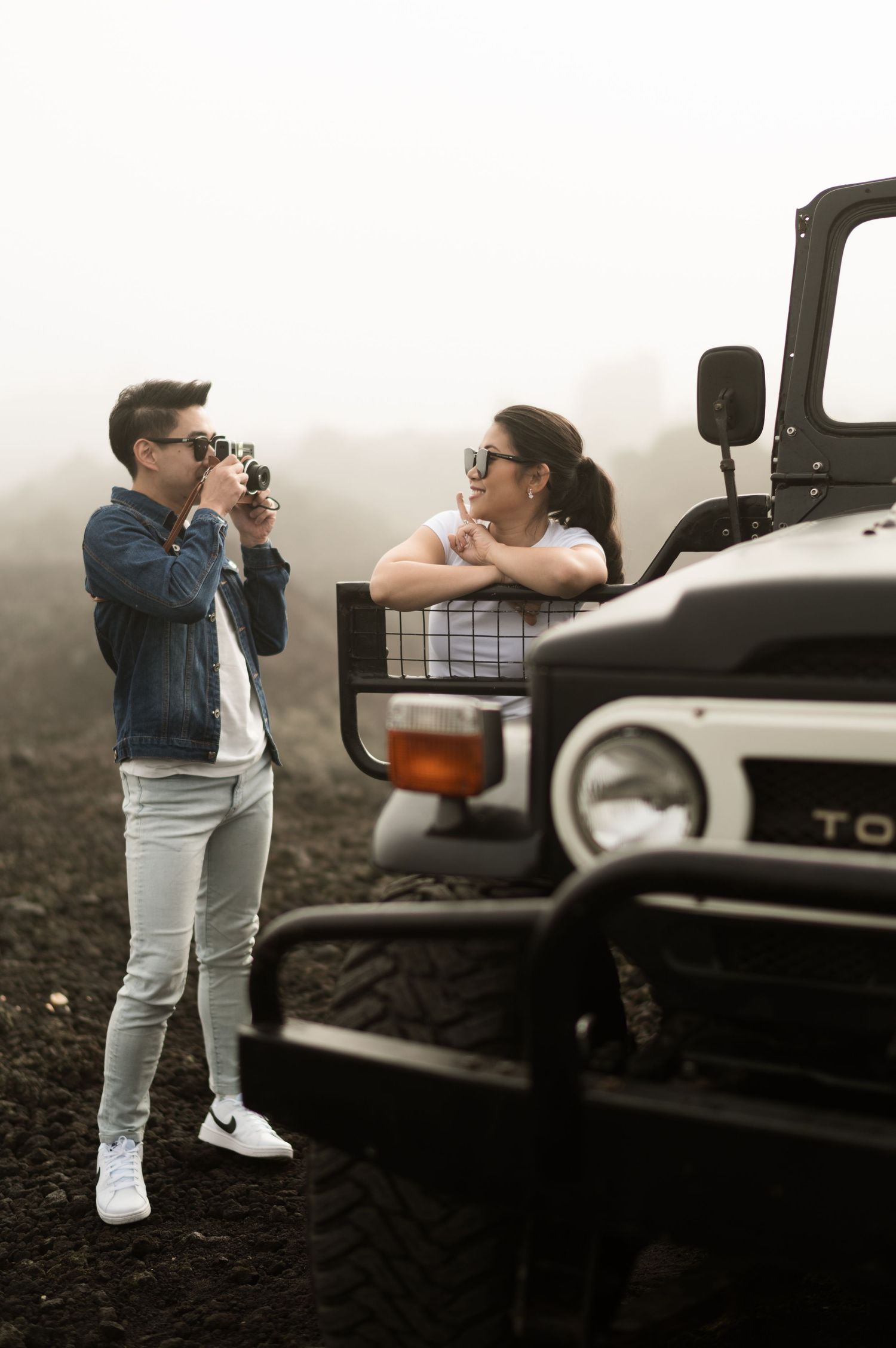 A man photographs a woman leaning on a vintage Toyota 4x4 in misty outdoor terrain.