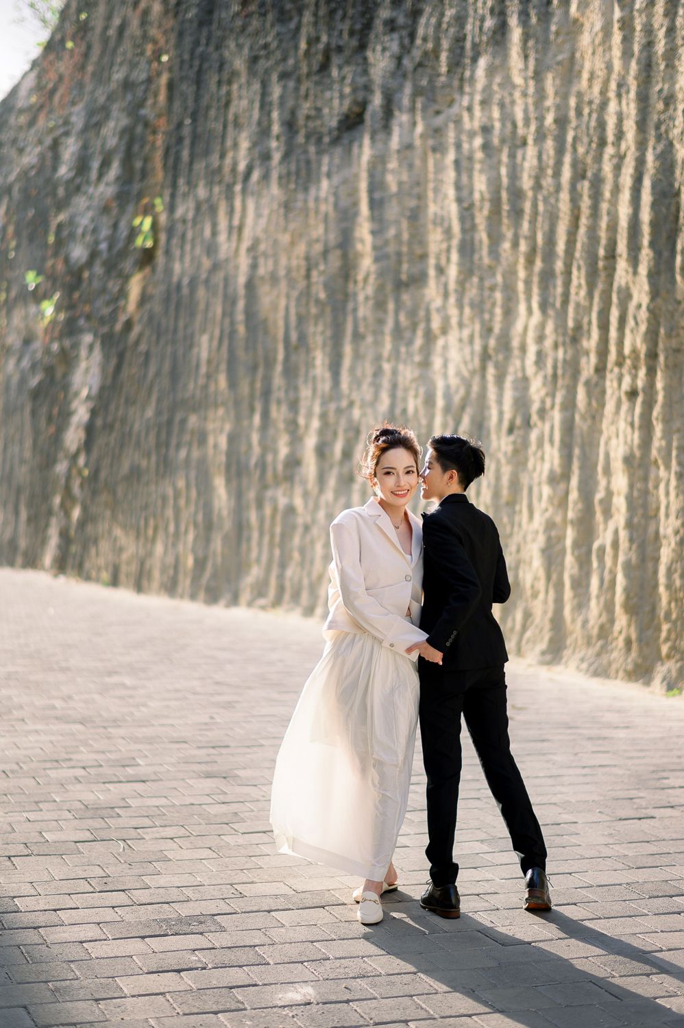 A couple poses romantically against a textured stone wall, the bride in white dress and groom in black suit sharing a tender kiss.