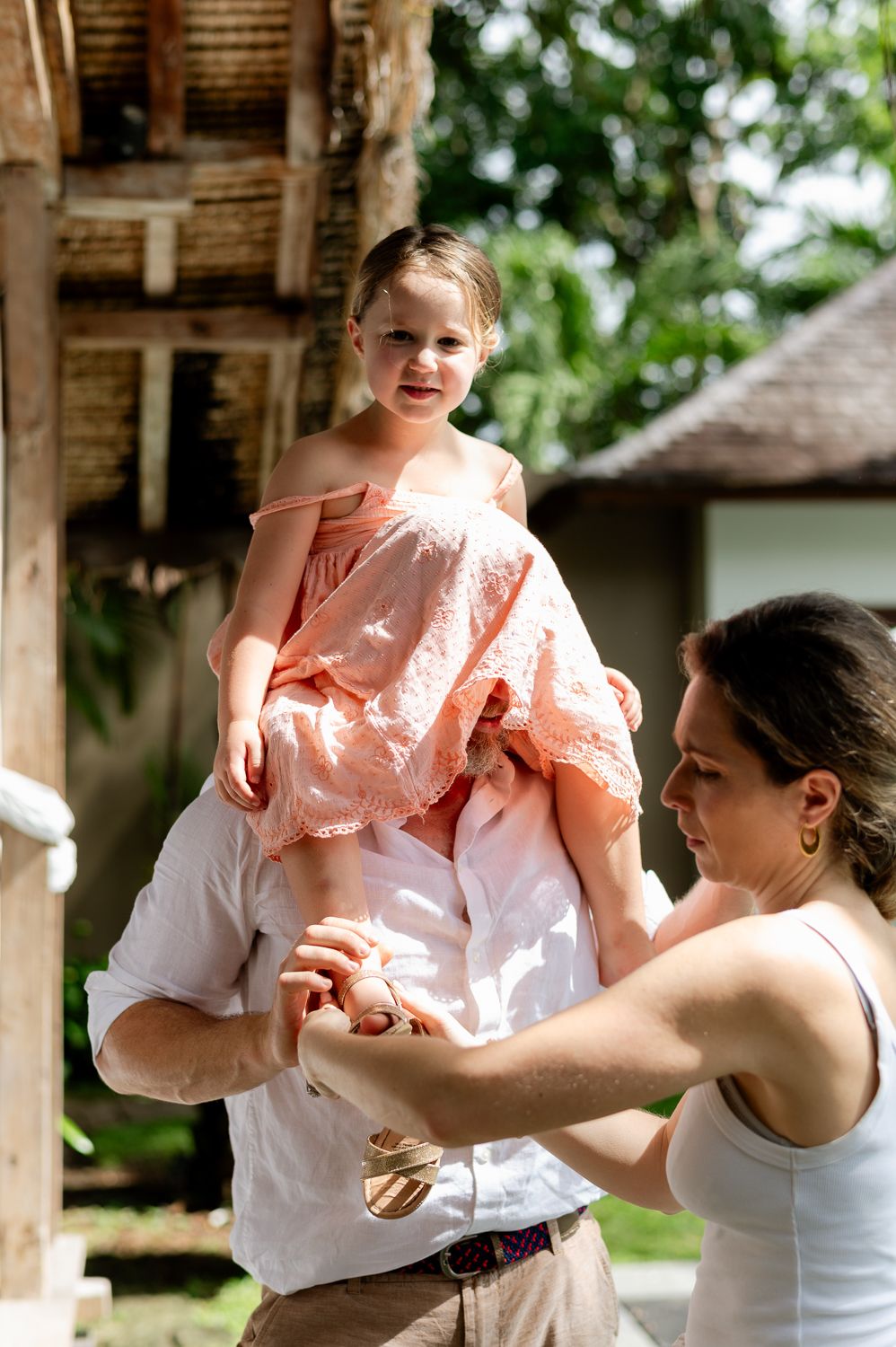 A man lifts a young girl in a pink dress onto his shoulders while a woman assists outdoors.