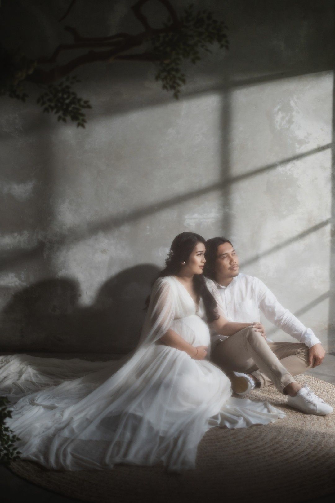Pregnant couple in white sitting together on floor against gray wall with dramatic light and shadow.