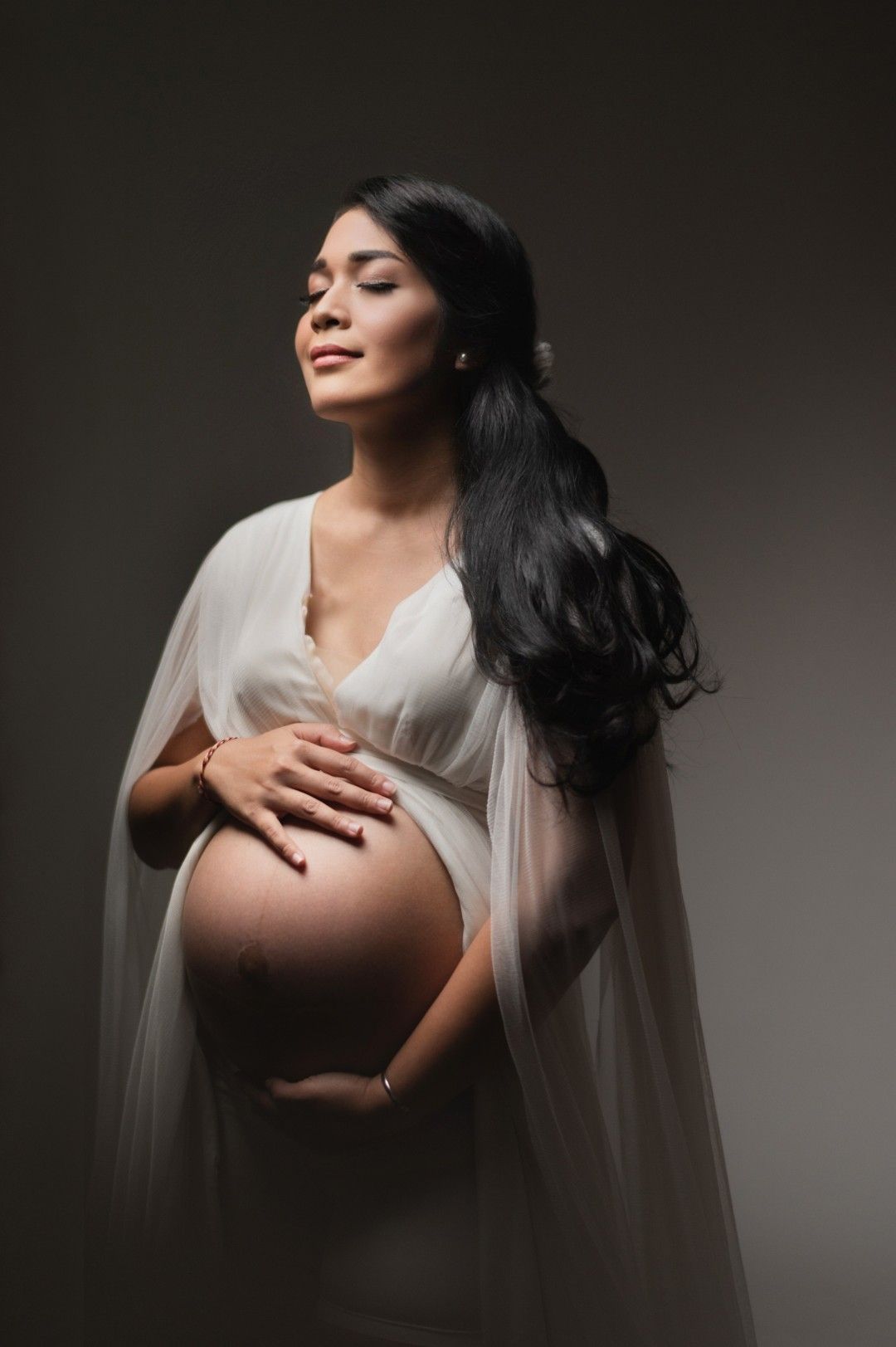 Elegant pregnant woman in white sheer gown cradling her baby bump in a soft-lit studio maternity portrait.