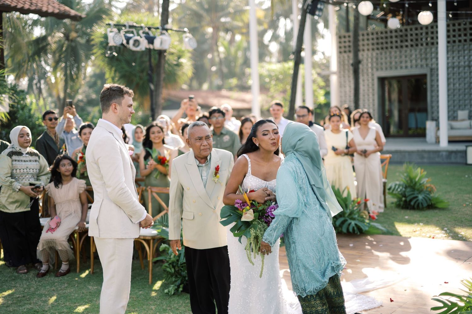 Bride walks down the aisle escorted by family at an outdoor tropical wedding ceremony.