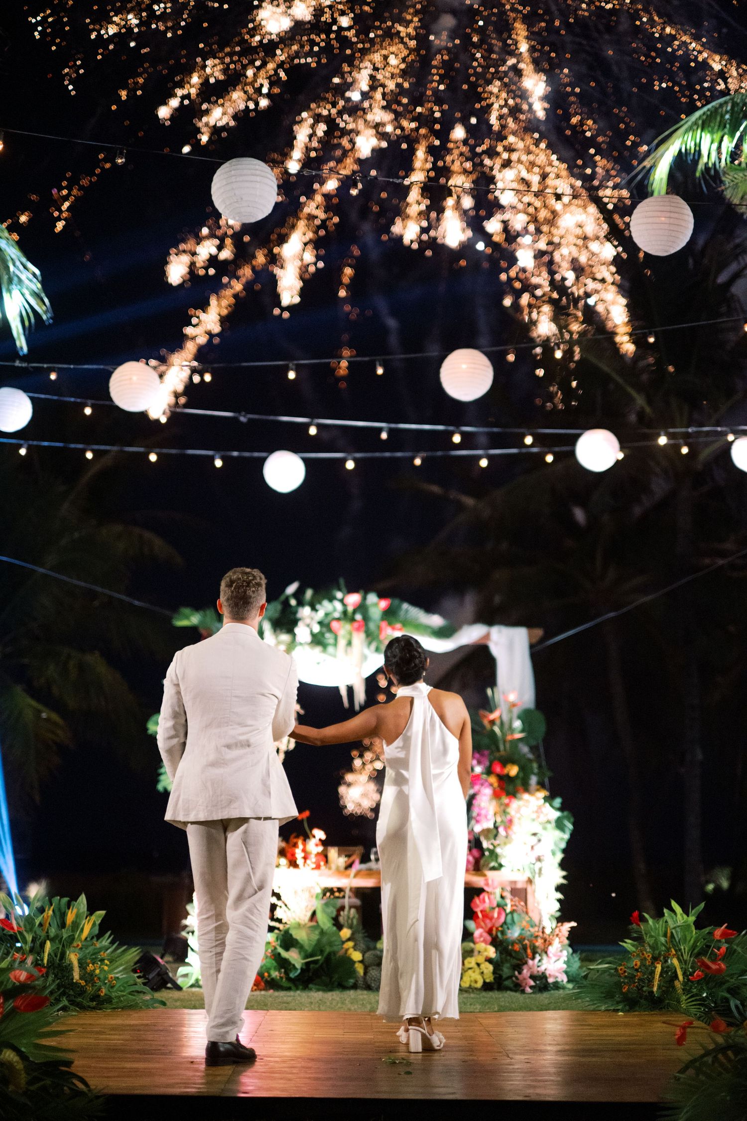 Bride and groom stand on dance floor watching fireworks at outdoor tropical wedding reception with string lights.
