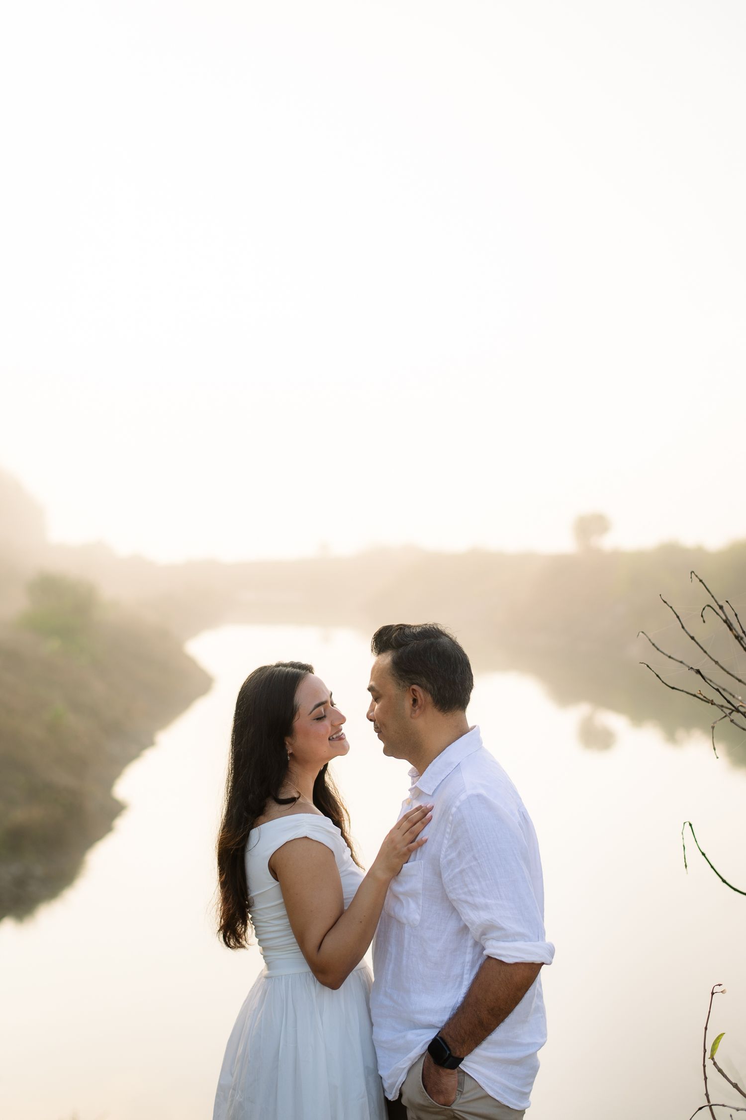 Couple in white outfits sharing a romantic moment by a misty river at golden hour.