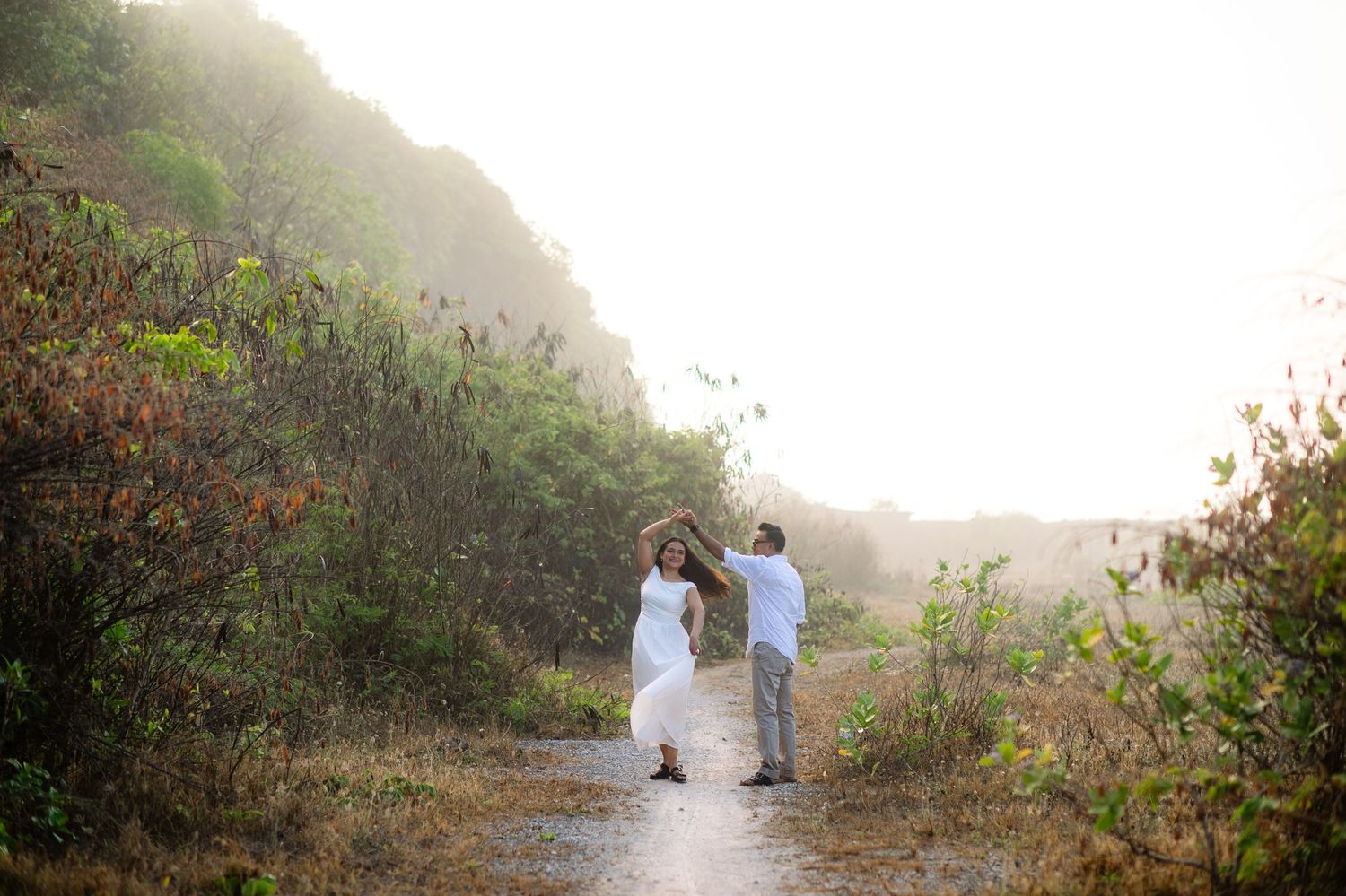 Couple dancing on a misty trail surrounded by lush greenery, woman in white dress, golden light in background.
