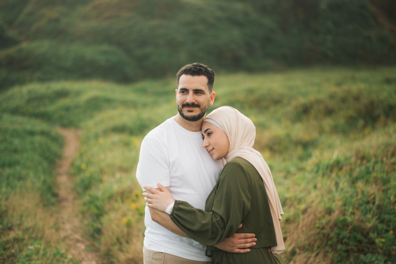 A couple embracing in a green field, woman wearing a hijab, man in white shirt, romantic outdoor portrait.