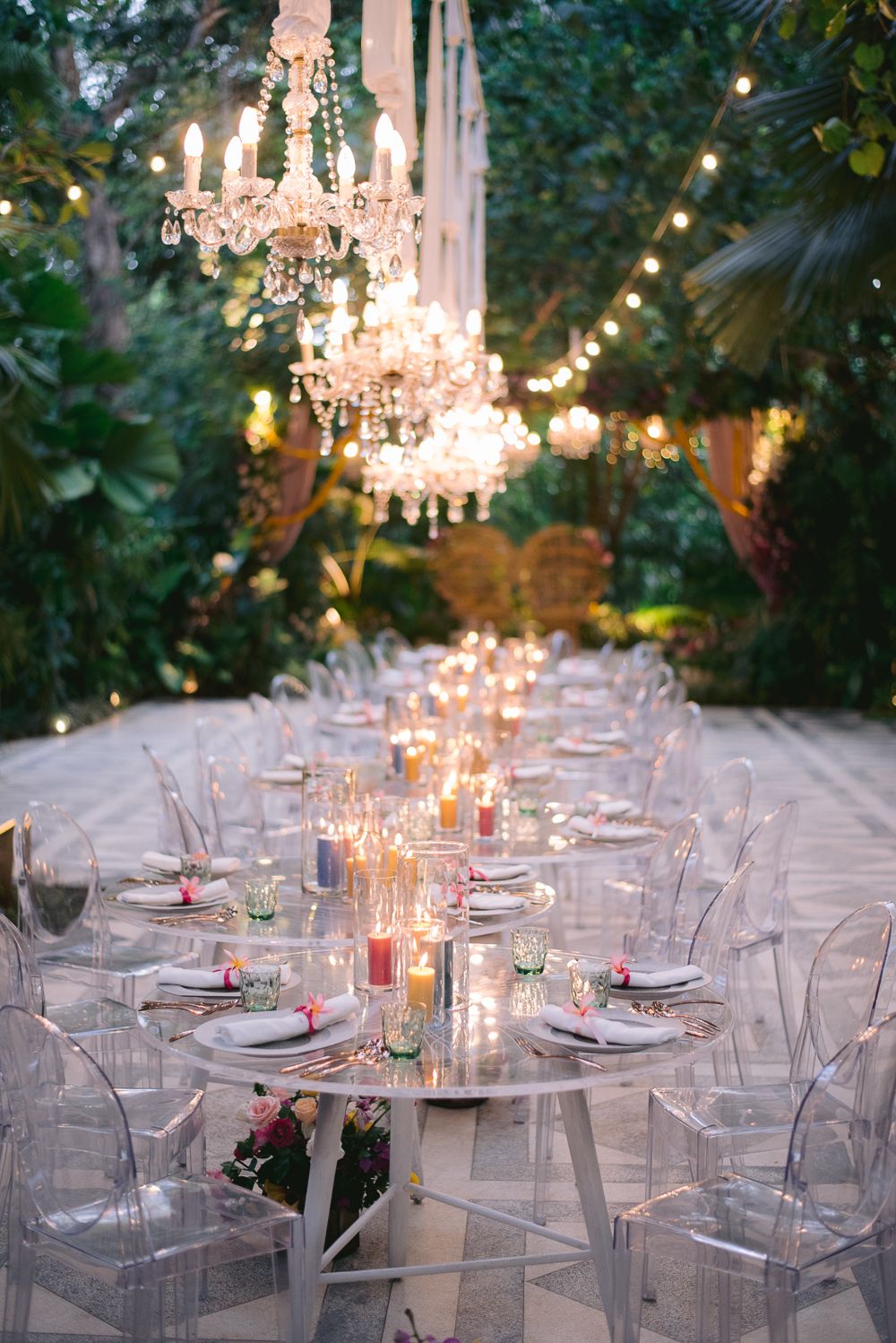 Elegant outdoor wedding reception with crystal chandeliers, ghost chairs, and candlelit long table surrounded by lush greenery.