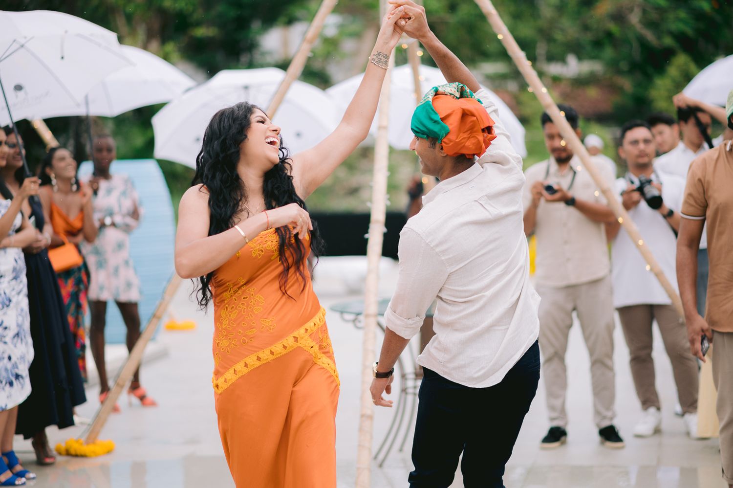 Couple dancing joyfully in orange outfits at an outdoor celebration with guests holding white umbrellas.