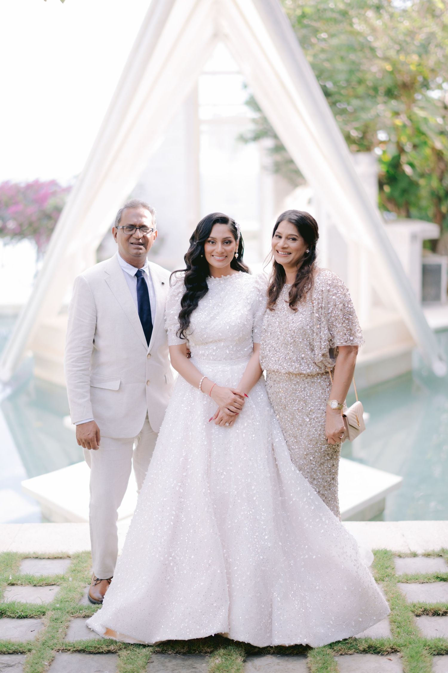 Bride in white sequin lehenga poses with parents at elegant outdoor wedding venue with pool backdrop.