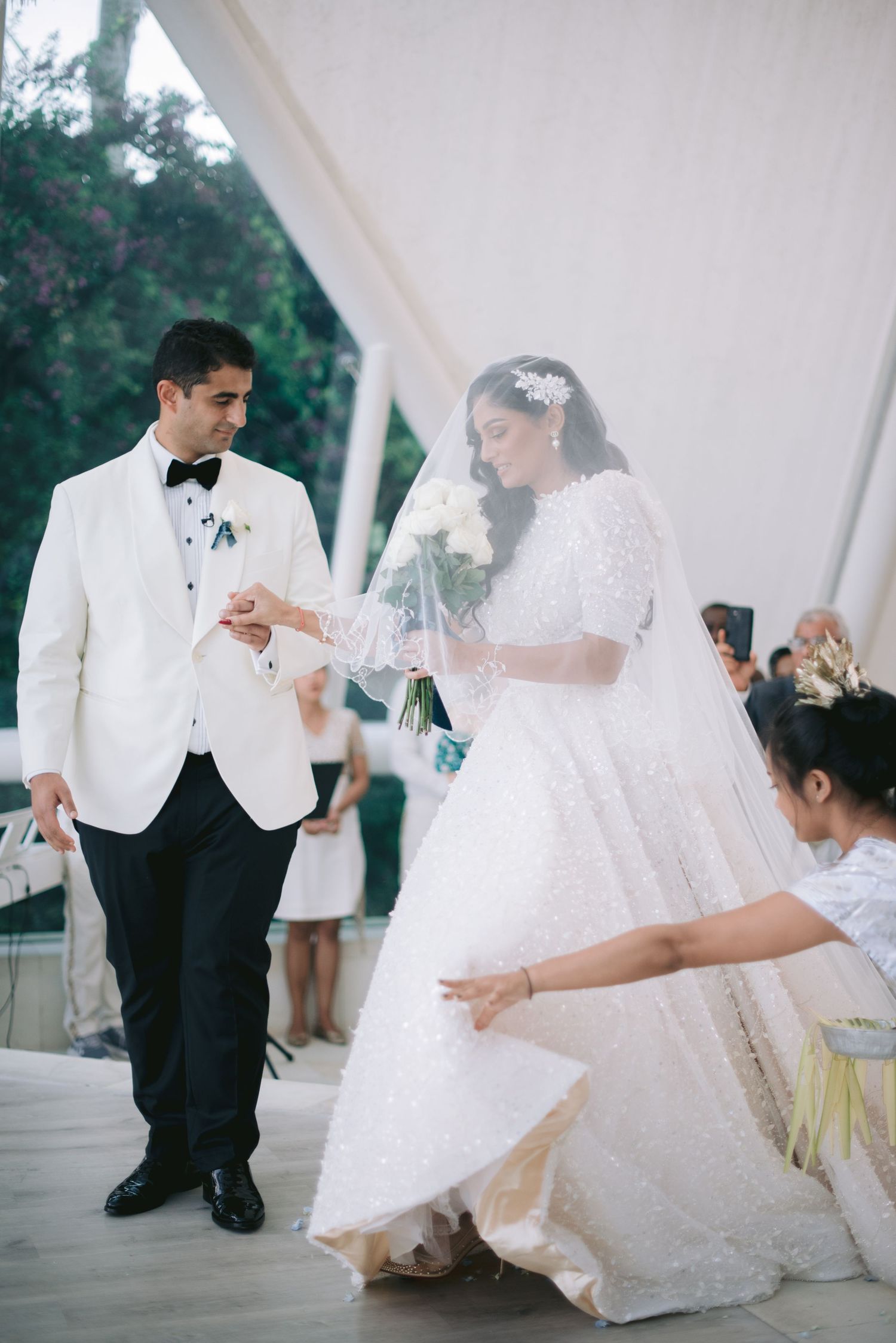 Bride in sequined gown and groom in white tuxedo exchange rings at elegant outdoor wedding ceremony.