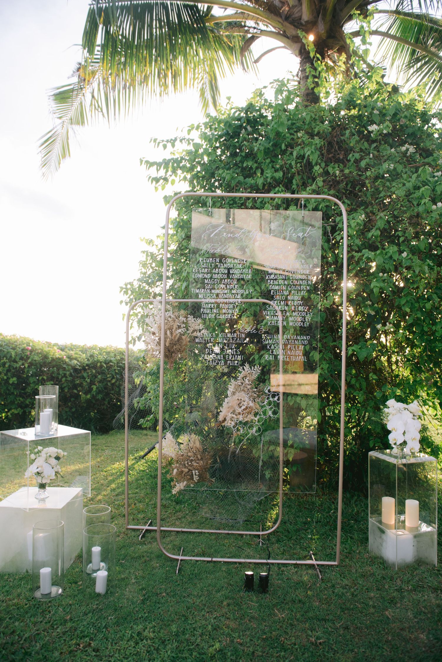 Elegant outdoor wedding seating chart displayed on acrylic panel in gold frame, surrounded by candles and greenery.