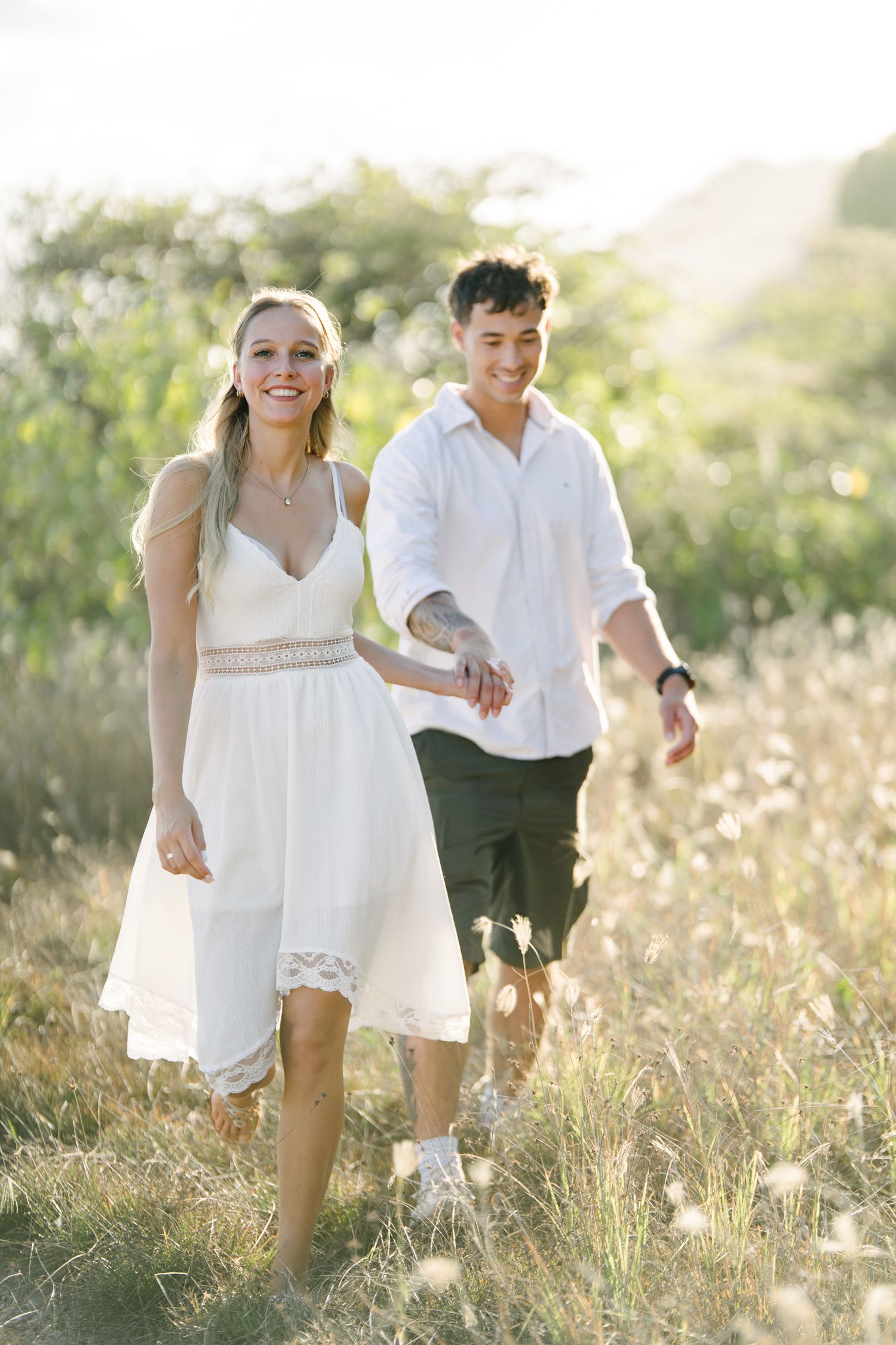 A smiling couple walks hand-in-hand through a sunny meadow, woman in white dress, man in white shirt and dark shorts.