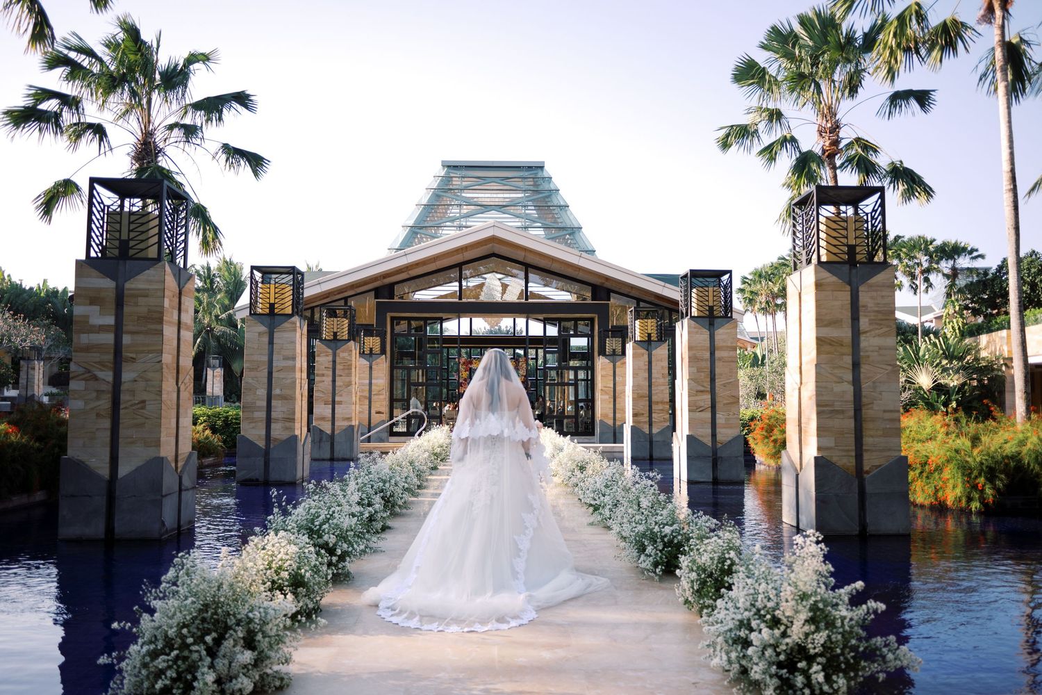 Bride in white gown and veil walks toward elegant tropical wedding venue flanked by palm trees and floral arrangements.