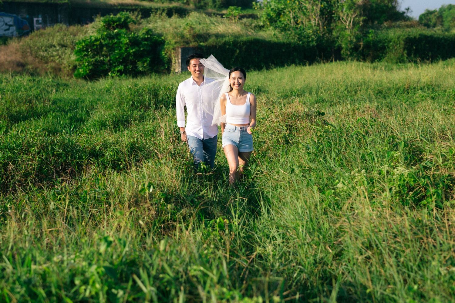 Happy couple walking through lush green meadow on a sunny day, man in white shirt and woman in denim shorts.