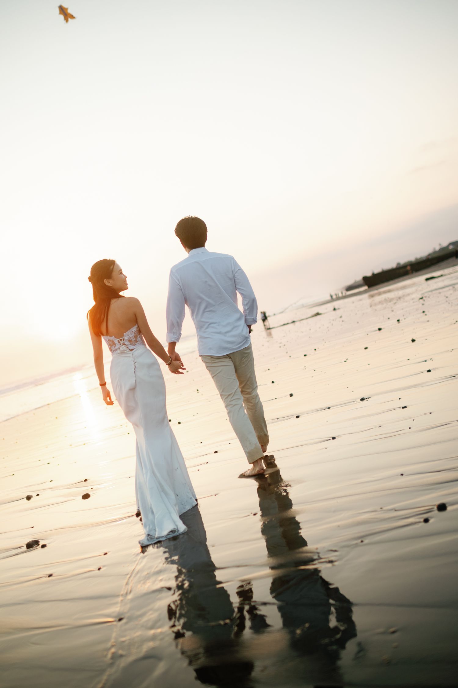 Couple walking hand in hand along a wet beach during golden sunset, woman in white dress.