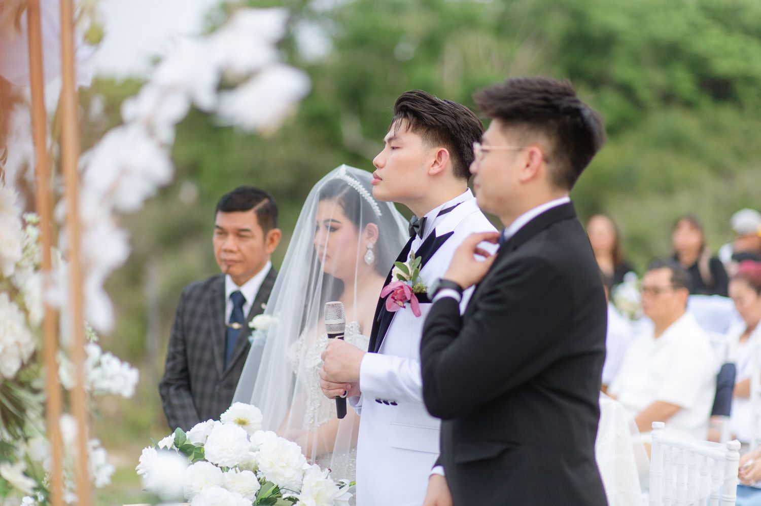 Outdoor wedding ceremony with bride in white gown and veil, groom in tuxedo, and guests gathered among floral decorations.