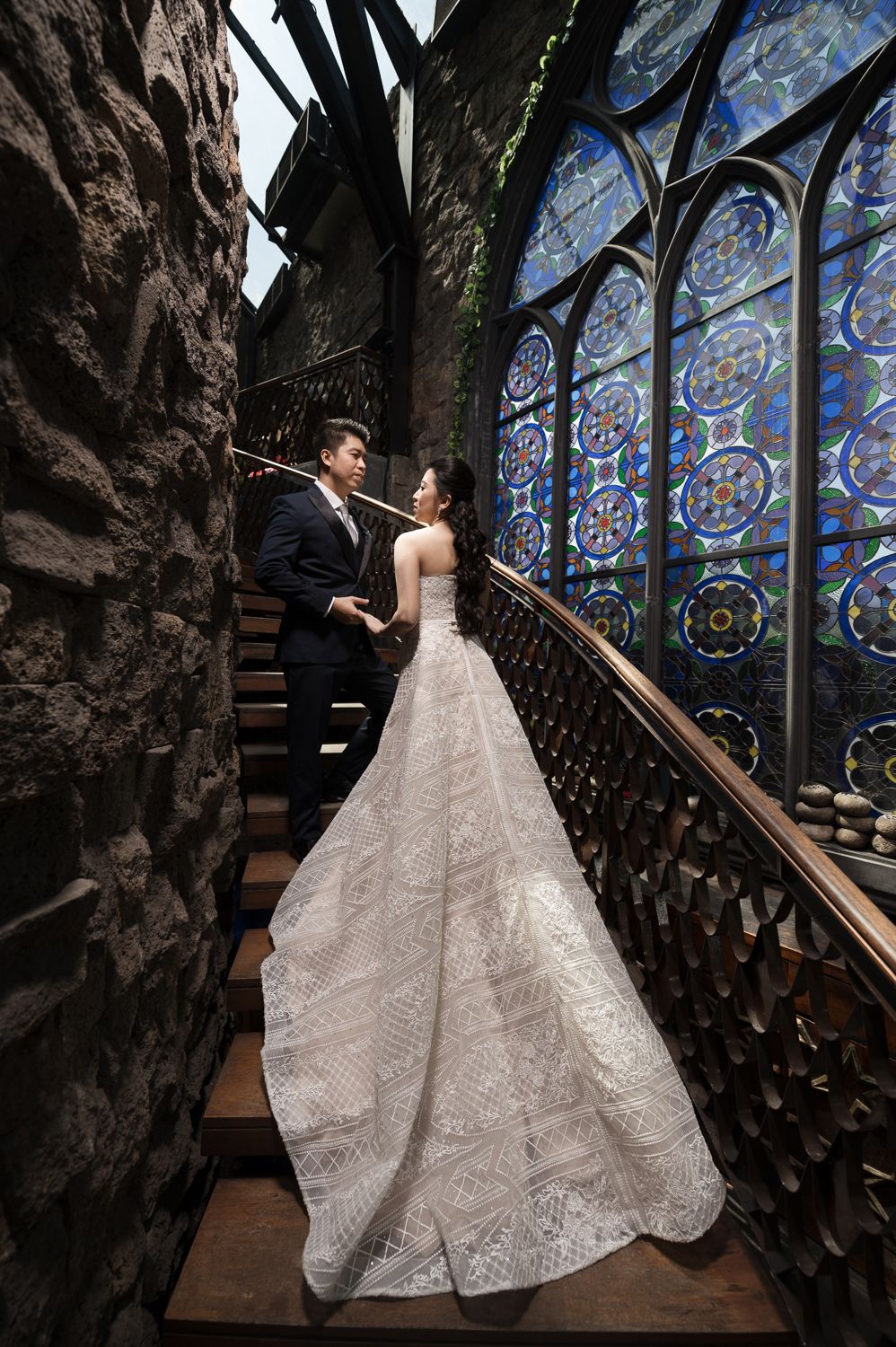 Couple in wedding attire poses on ornate staircase with stunning stained glass windows in gothic architecture.