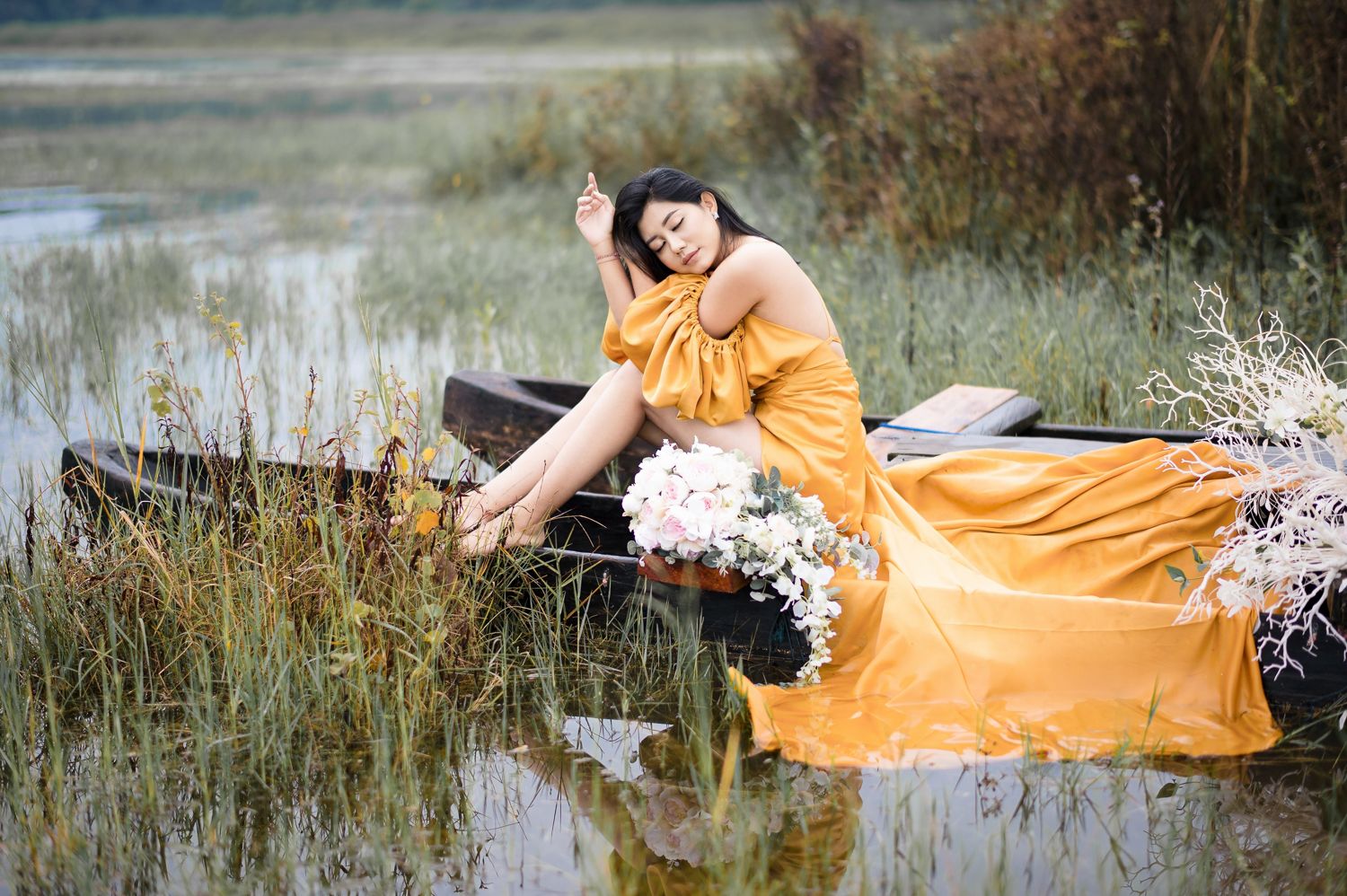 Woman in golden yellow off-shoulder gown sitting gracefully in a wooden boat surrounded by wildgrass and white flowers.