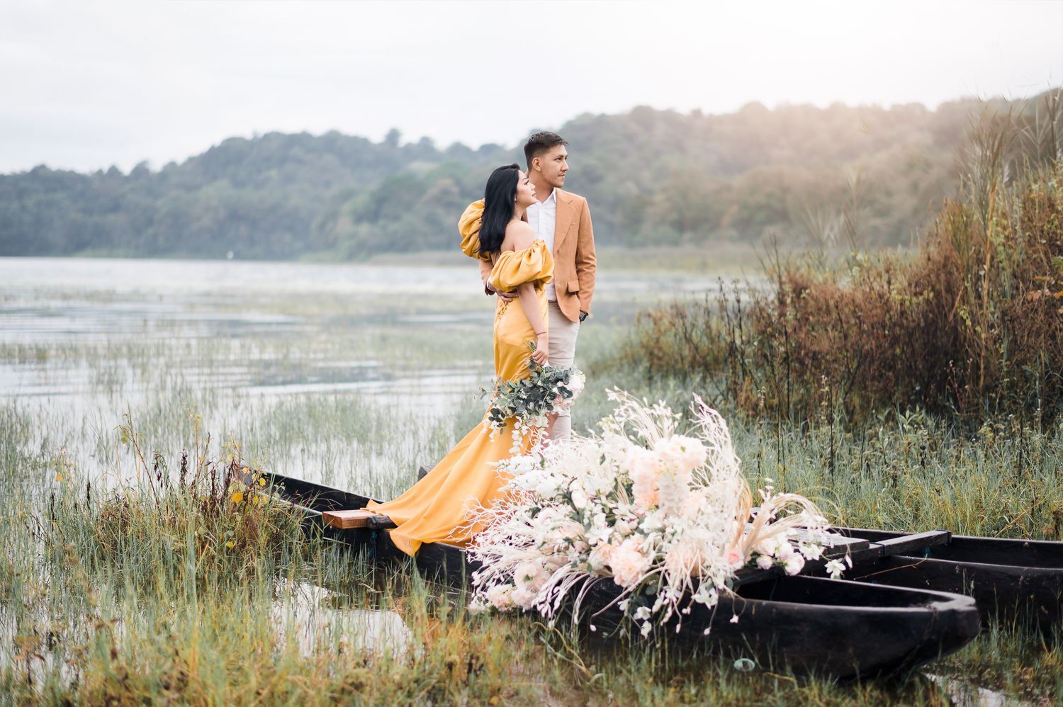 Couple in elegant attire standing in a rustic boat adorned with flowers by a serene lake surrounded by lush greenery.