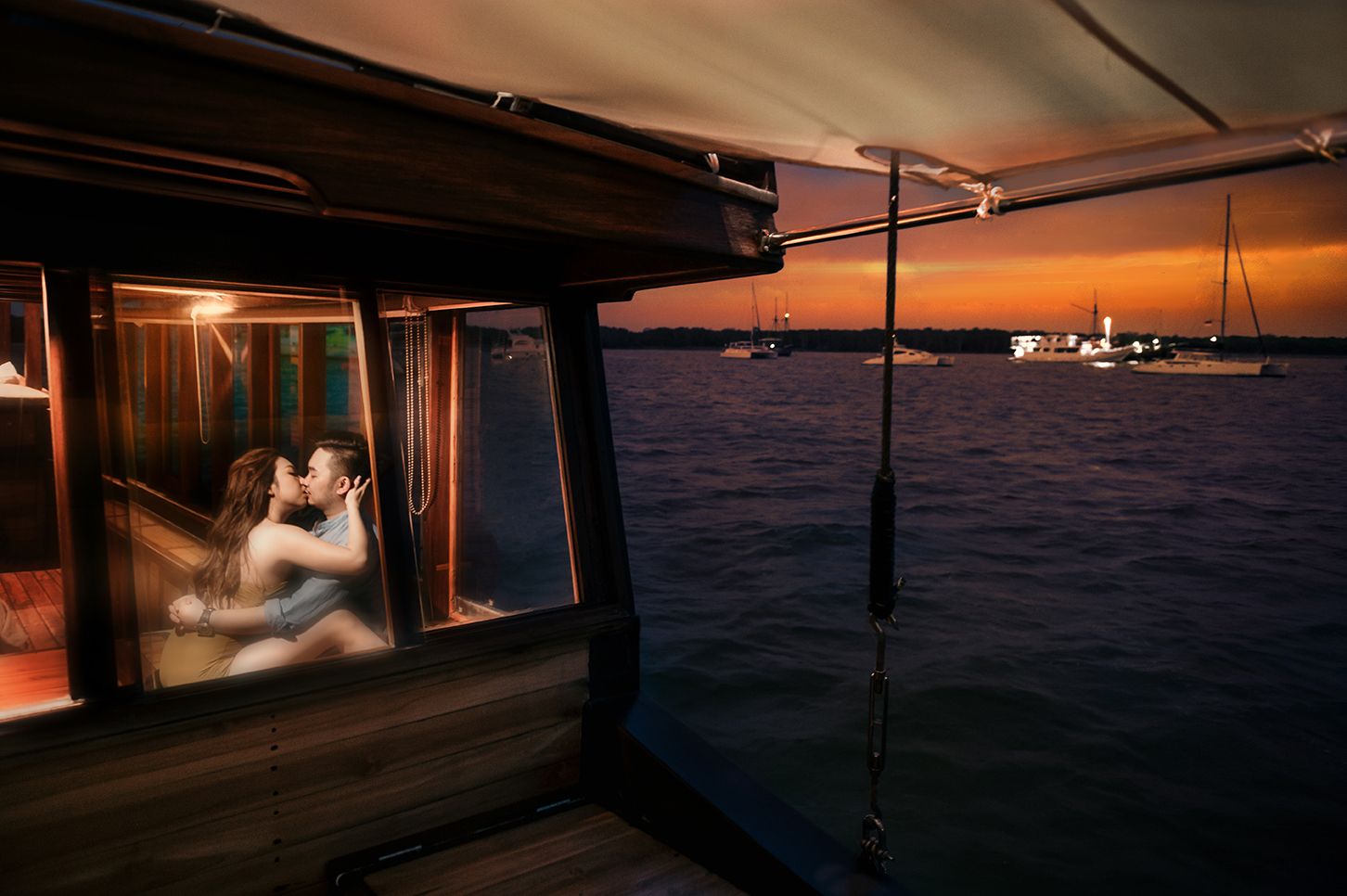 Couple sharing a romantic kiss inside a boat cabin at sunset over calm ocean waters.