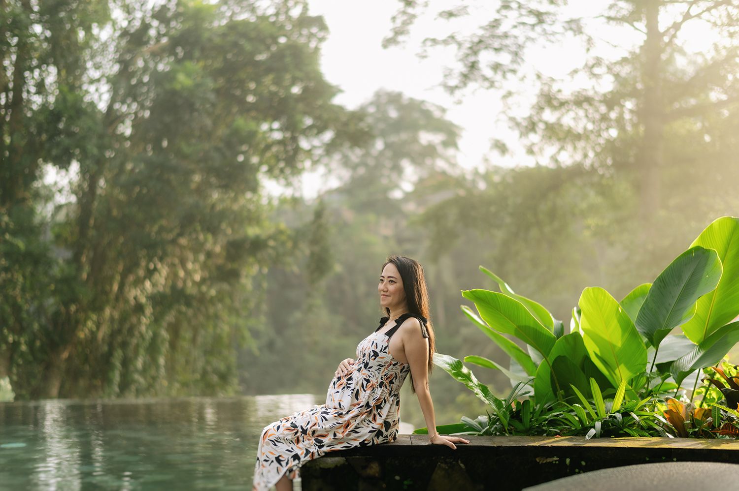 Pregnant woman in floral dress sitting by a lush tropical infinity pool surrounded by green plants and misty jungle Maya Ubud Resort and Sp.