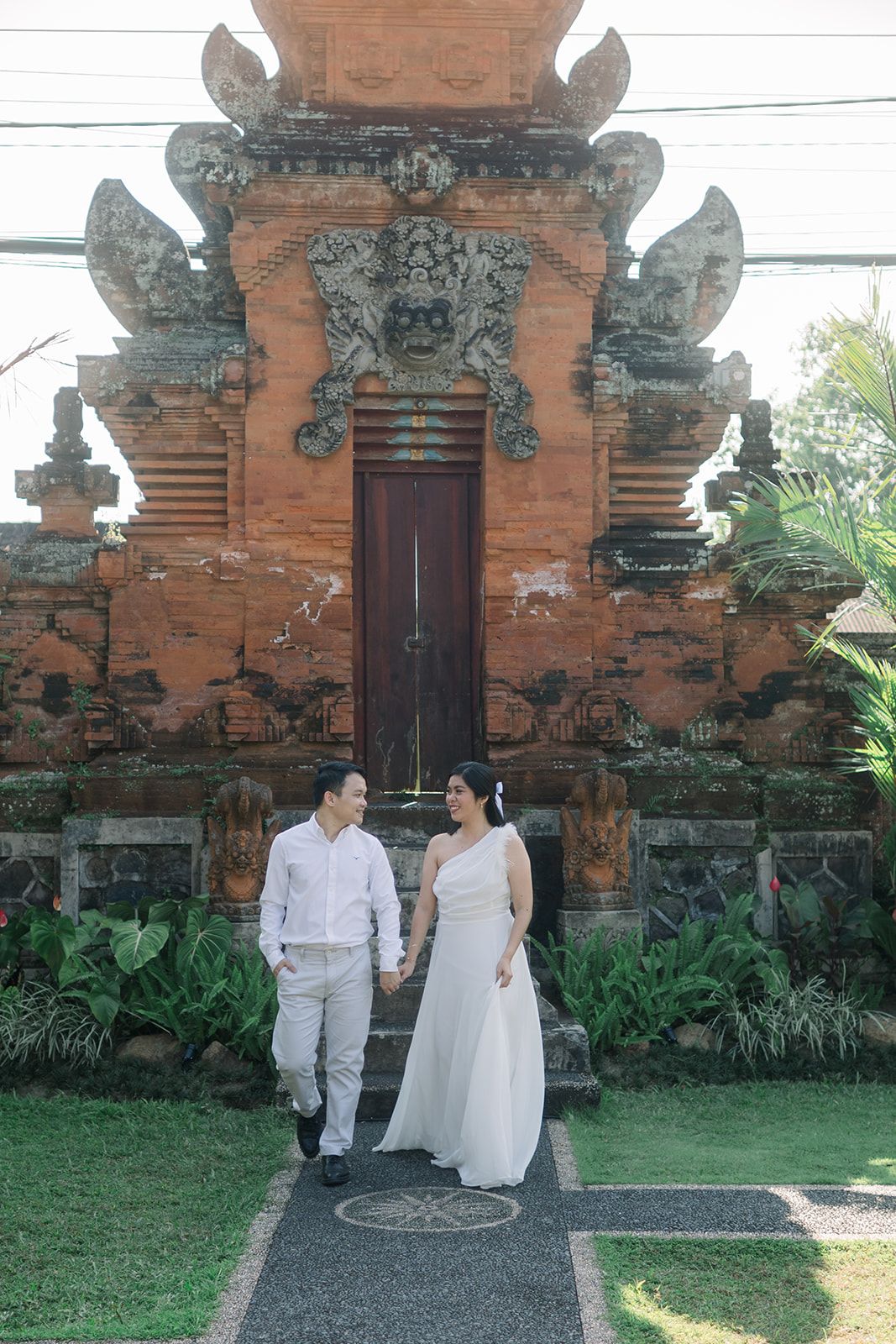 Couple holding hands walks before an ornate traditional Balinese temple gate with intricate stone carvings.