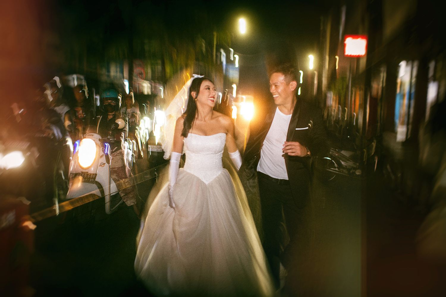 A joyful bride and groom walk together at night, surrounded by bright lights, as she wears a stunning white ball gown.