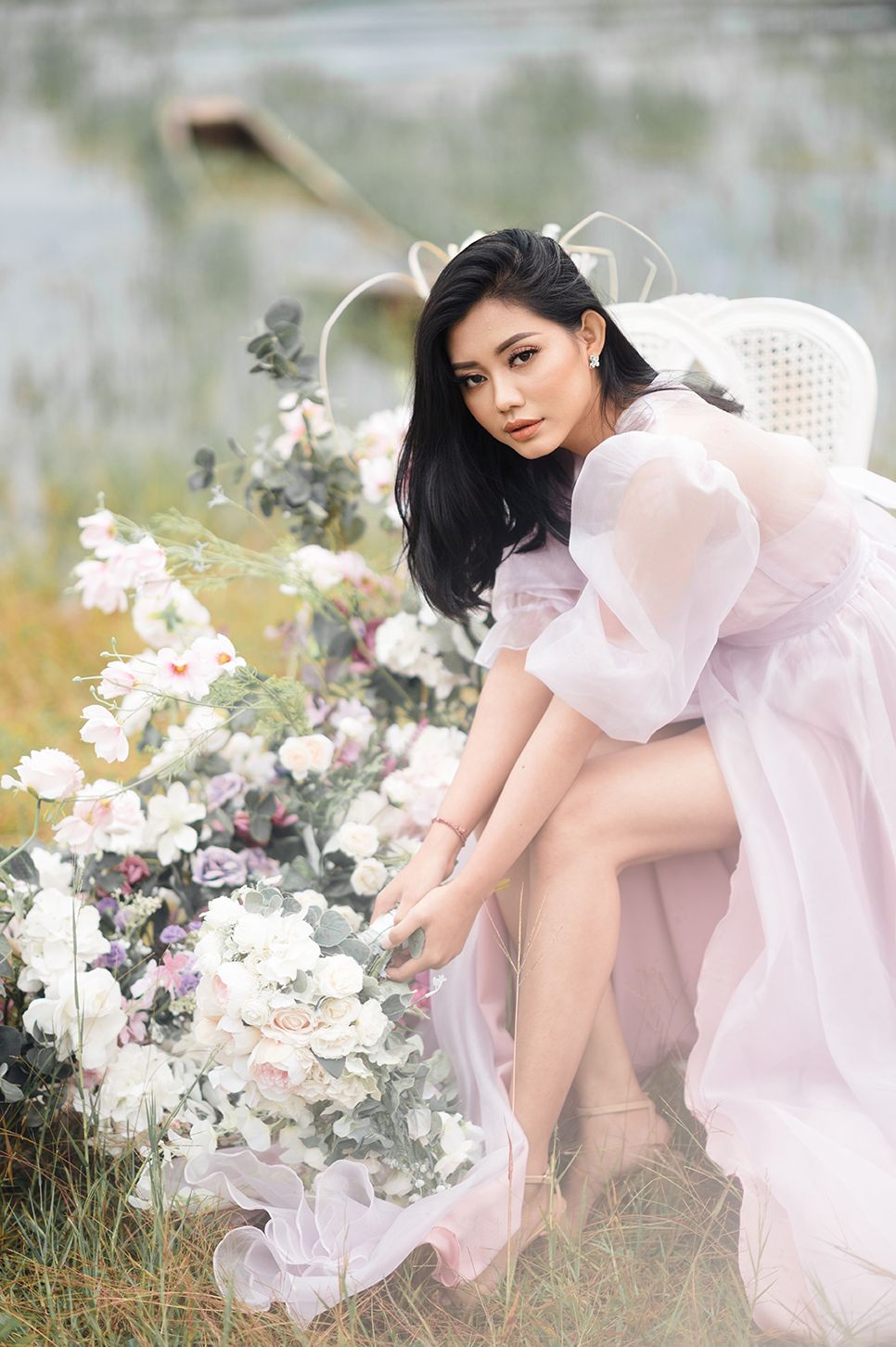 A woman in a pink tulle gown poses gracefully among white roses outdoors, with long black hair and elegant styling.