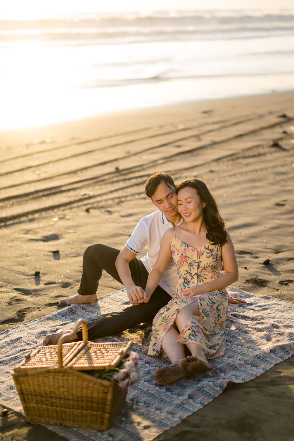 Couple sitting on a blanket at the beach during golden hour with a picnic basket, woman in floral dress cradling baby bump.