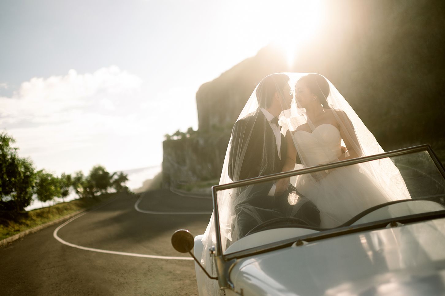Bride and groom kissing in a vintage convertible car on a scenic road, bathed in golden sunset light.