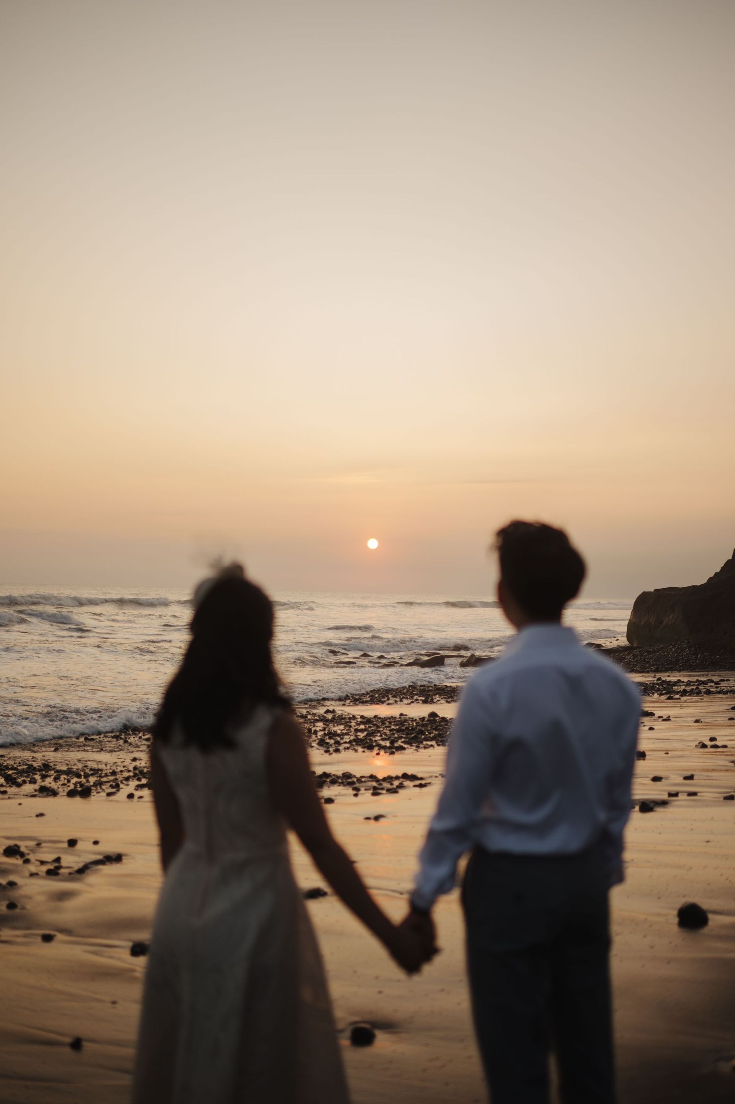 Couple in wedding attire holds hands on a sandy beach watching a romantic golden sunset over ocean waves.