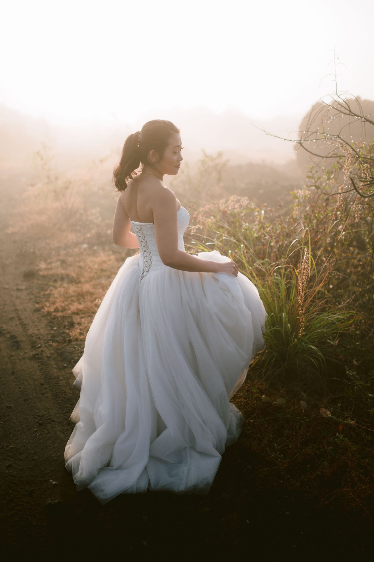 Bride in a flowing white tulle ball gown stands on a misty outdoor path, glowing in soft golden light.