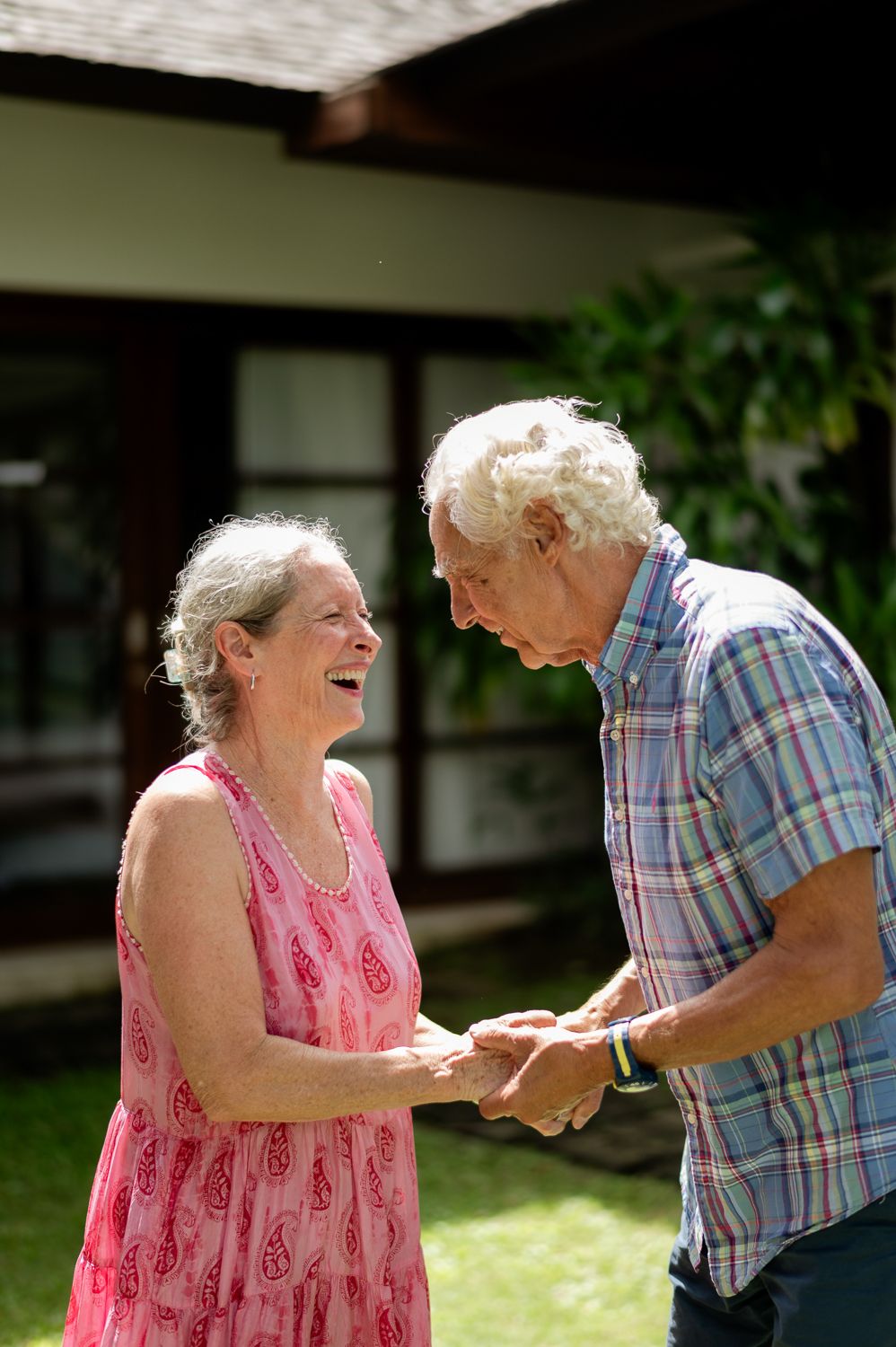 Happy senior couple holding hands and laughing together outdoors in a sunny garden near a house.
