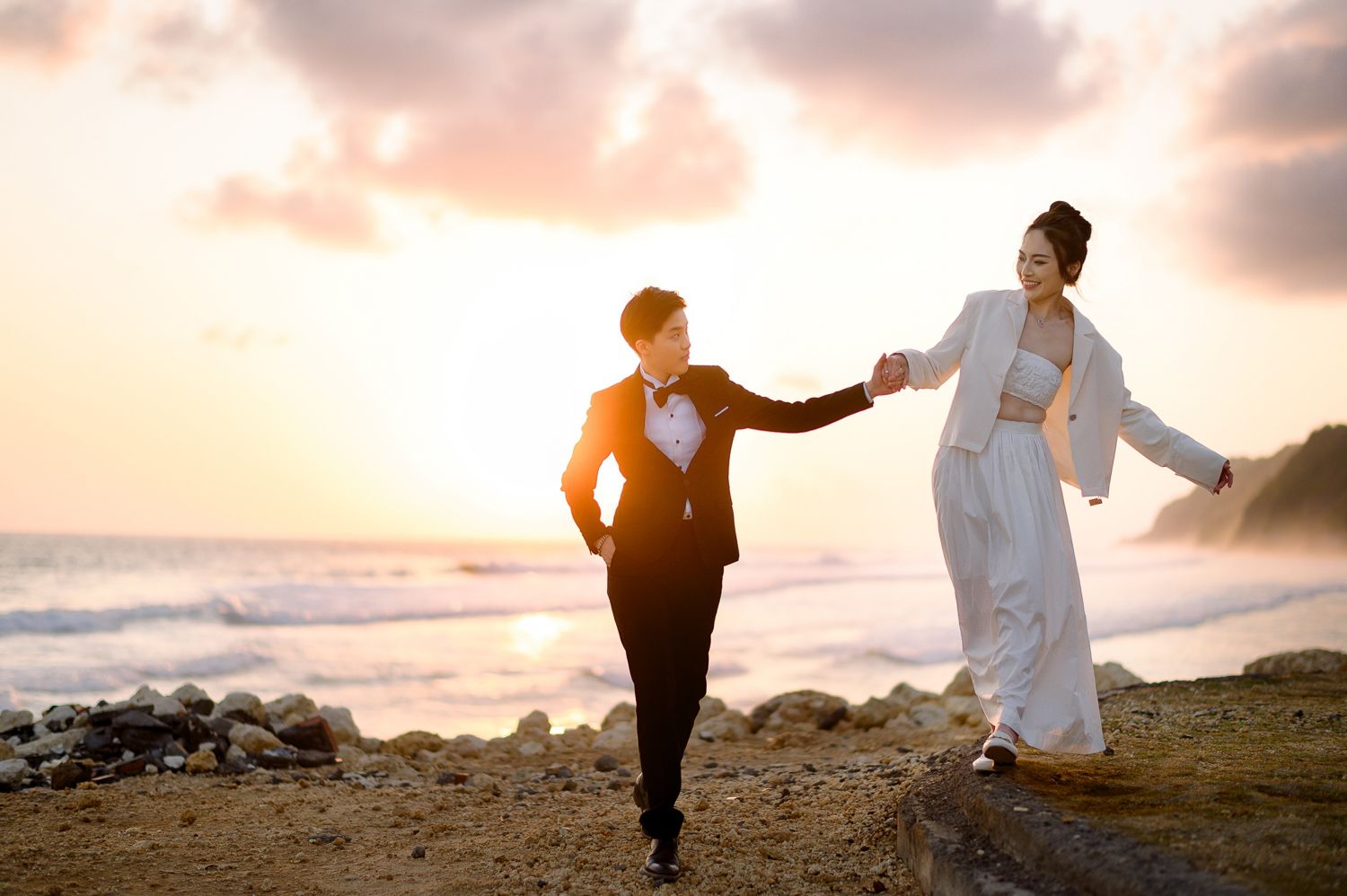 Couple in wedding attire holding hands on a beach at sunset, with ocean waves and colorful sky behind them.