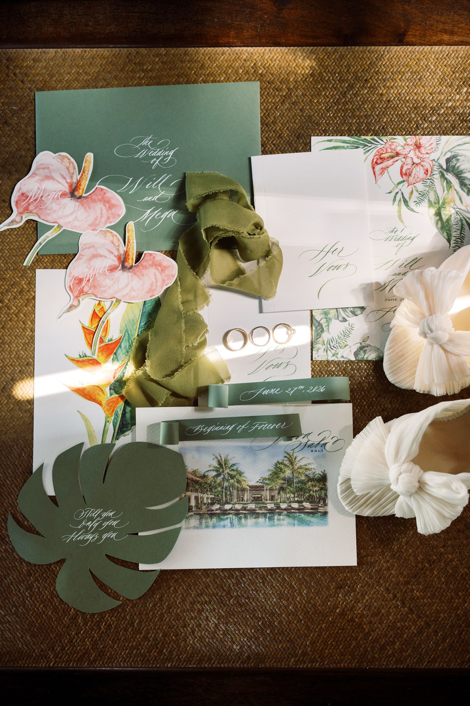 Tropical wedding stationery flatlay with floral invitations, monstera leaves, rings, and bridal shoes on a textured surface.