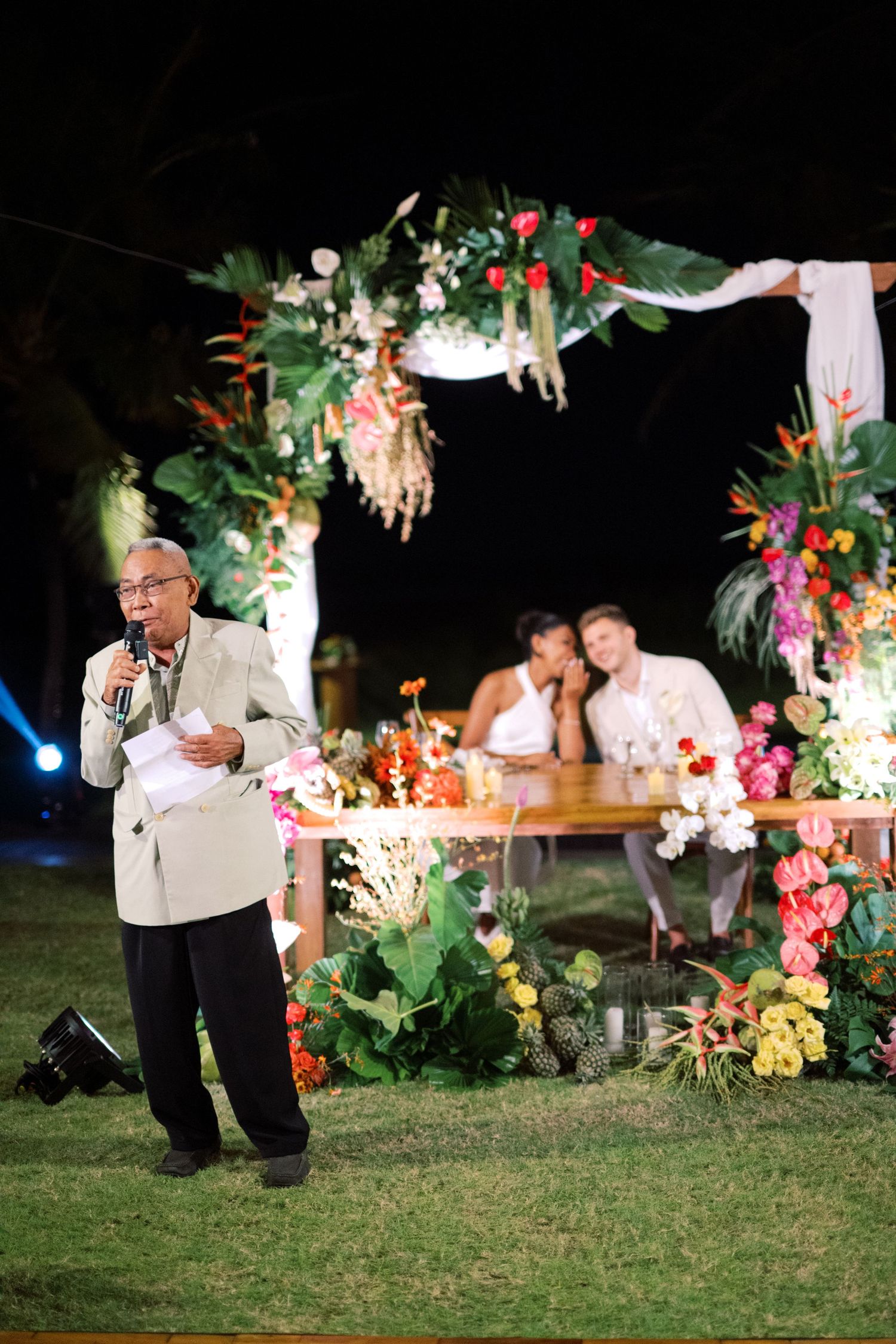 Elderly man gives speech at outdoor tropical wedding reception while couple listens.