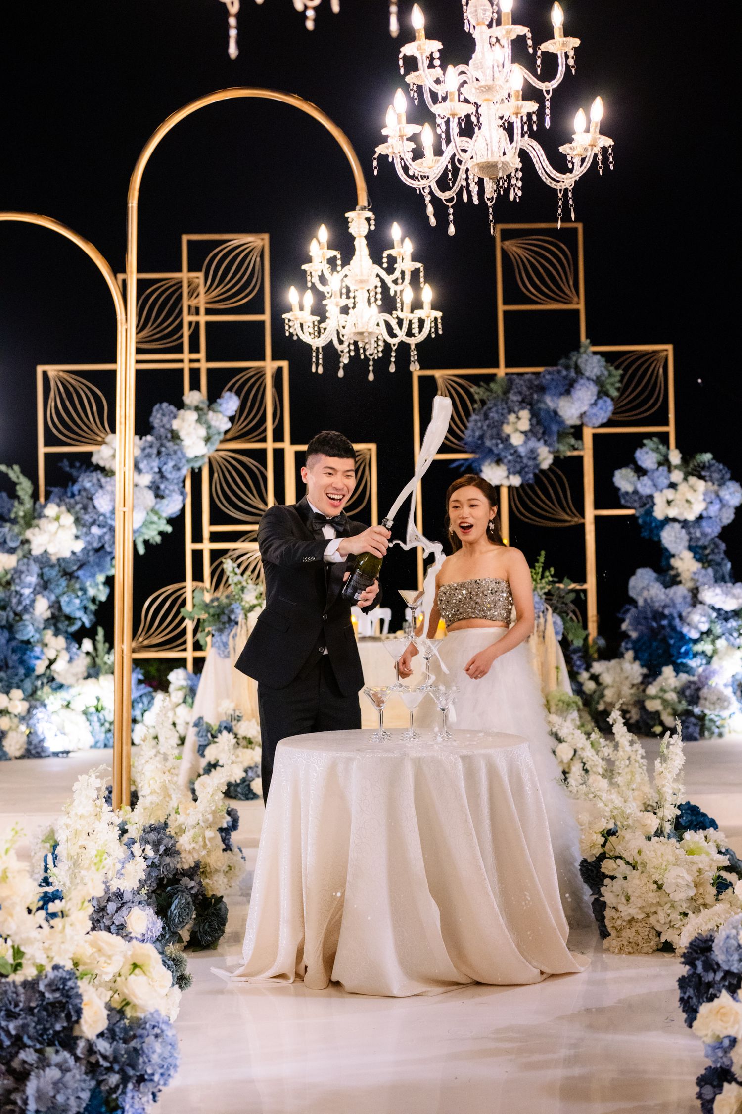 A smiling groom and bride pour champagne at an elegant wedding reception with gold arches, crystal chandeliers, and blue and white flowers.