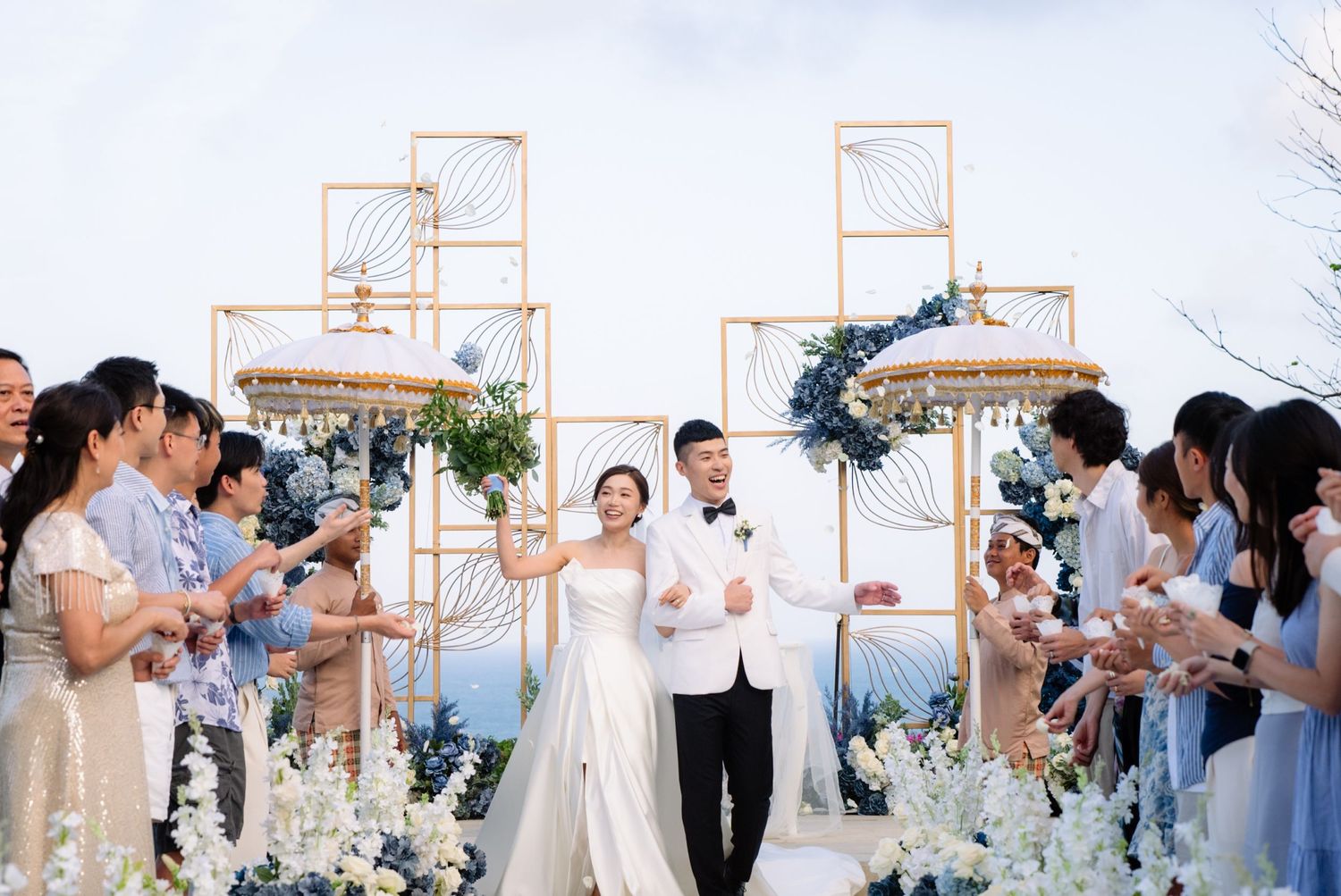 Bride in a white gown and groom in a white tuxedo walk down the aisle at an outdoor wedding as guests cheer among blue and white flowers.