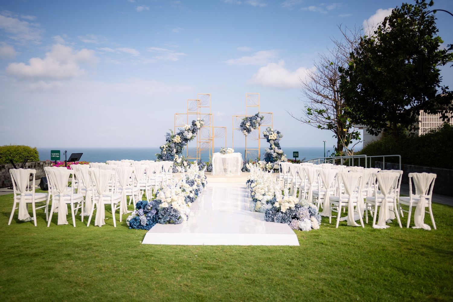 Outdoor wedding ceremony setup with white chairs, blue and white floral arrangements, and a floral arch overlooking the ocean.