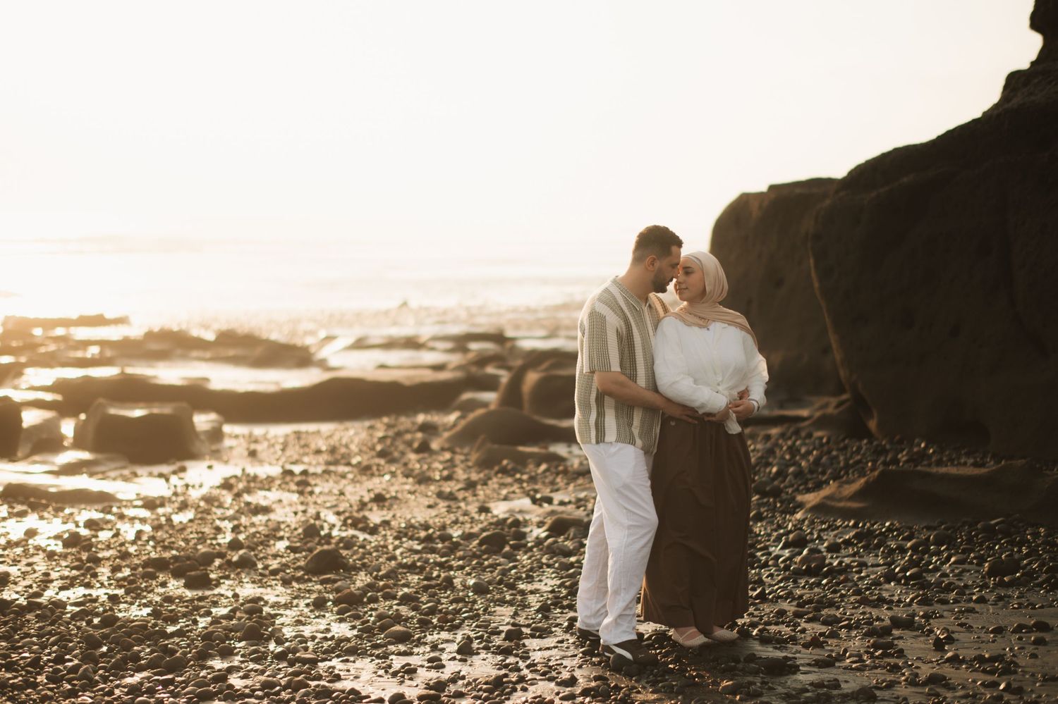 Couple sharing a romantic kiss on a rocky beach at golden sunset, dressed in elegant casual attire.
