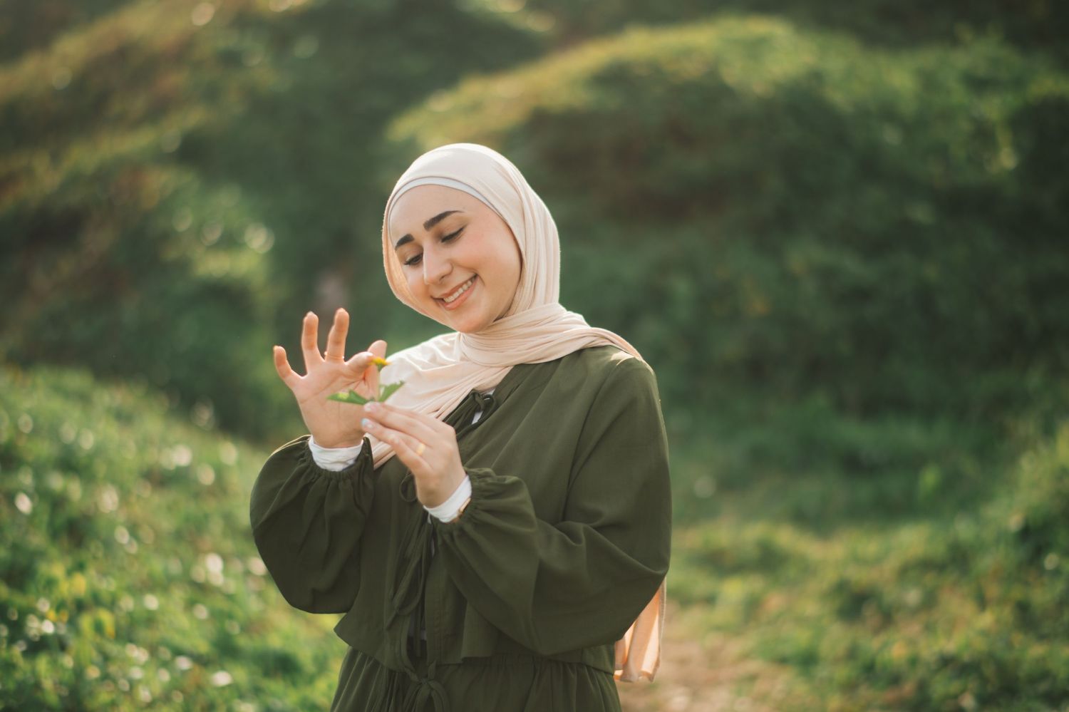 A smiling woman in a green dress and beige hijab holds a small flower outdoors in a lush green setting.