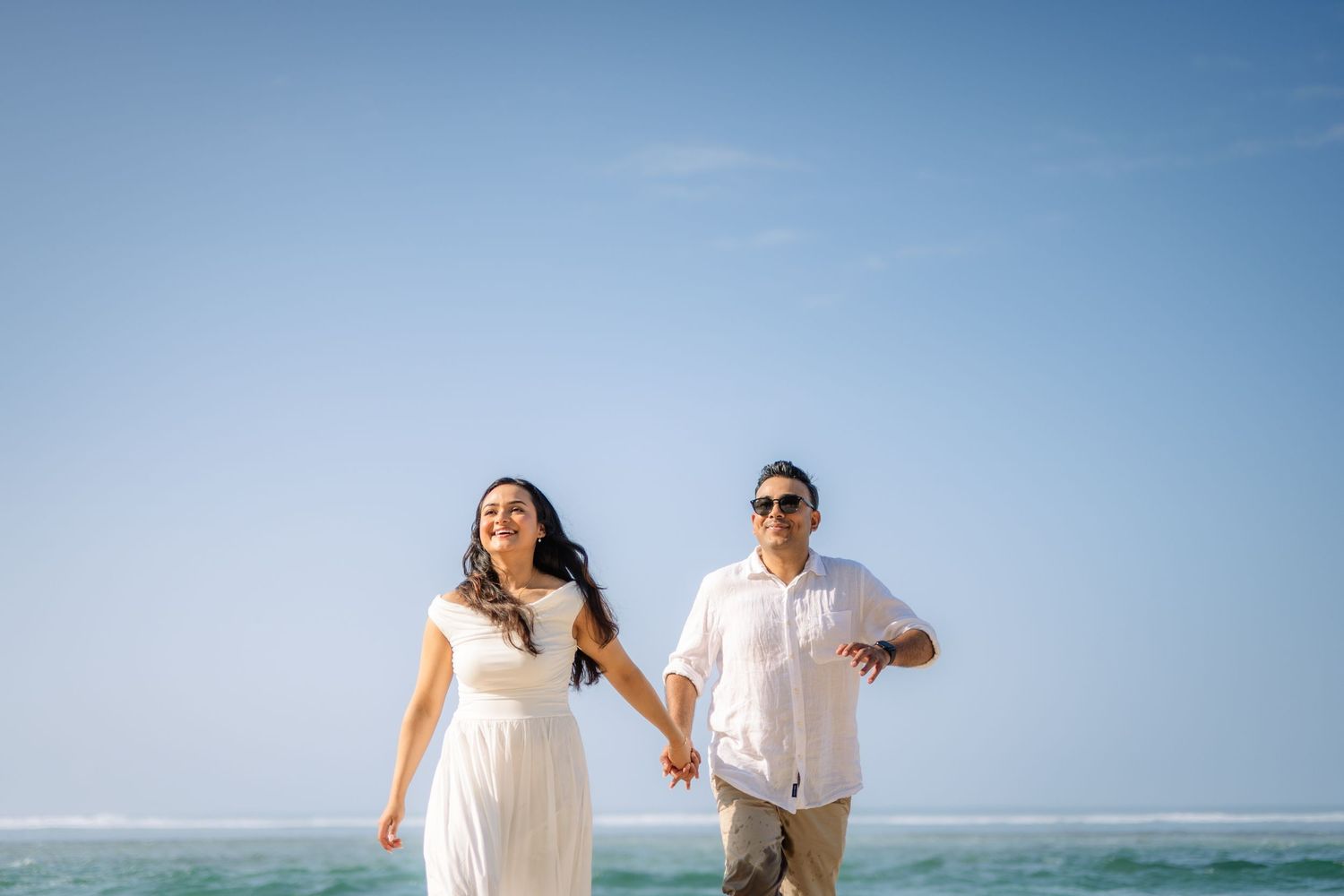 Happy couple in white outfits holding hands and walking on a sunny beach with blue ocean water behind them.