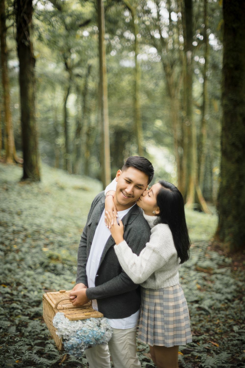 Couple sharing a romantic moment in a lush green forest, man holding a wicker picnic basket with blue flowers.
