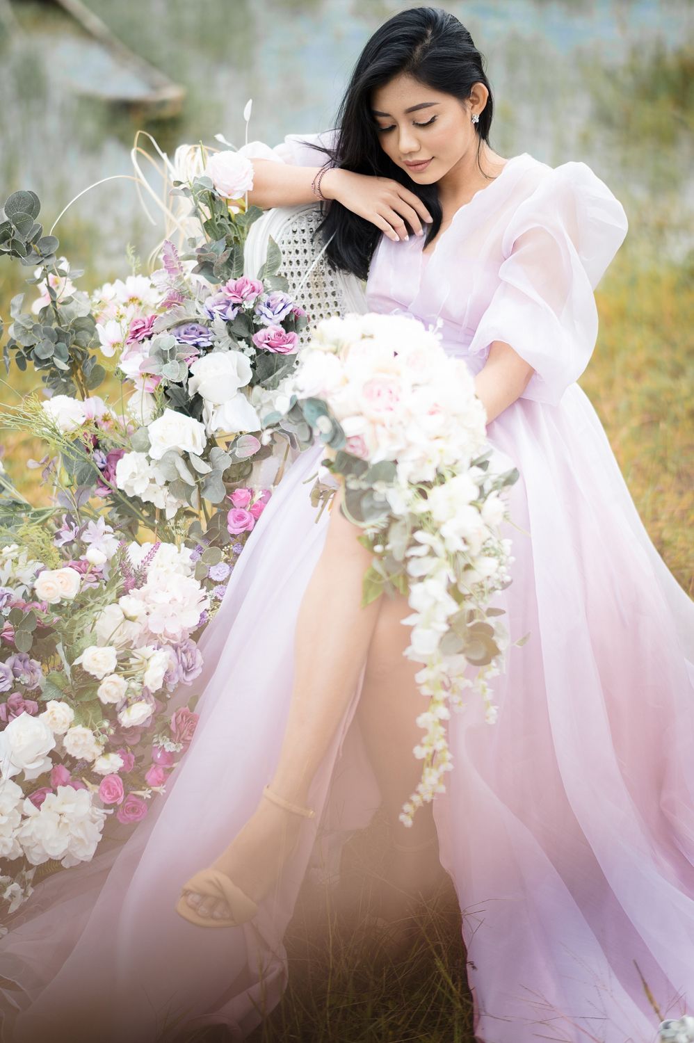 A bride in a flowing pink tulle gown holds a cascading white floral bouquet outdoors among blooming flowers.