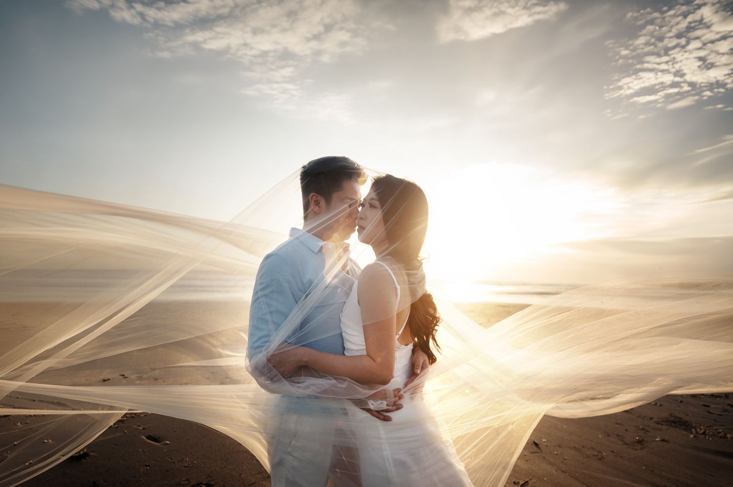 Couple sharing a romantic kiss on sandy desert dunes, bride's veil flowing in wind against golden sunset sky.
